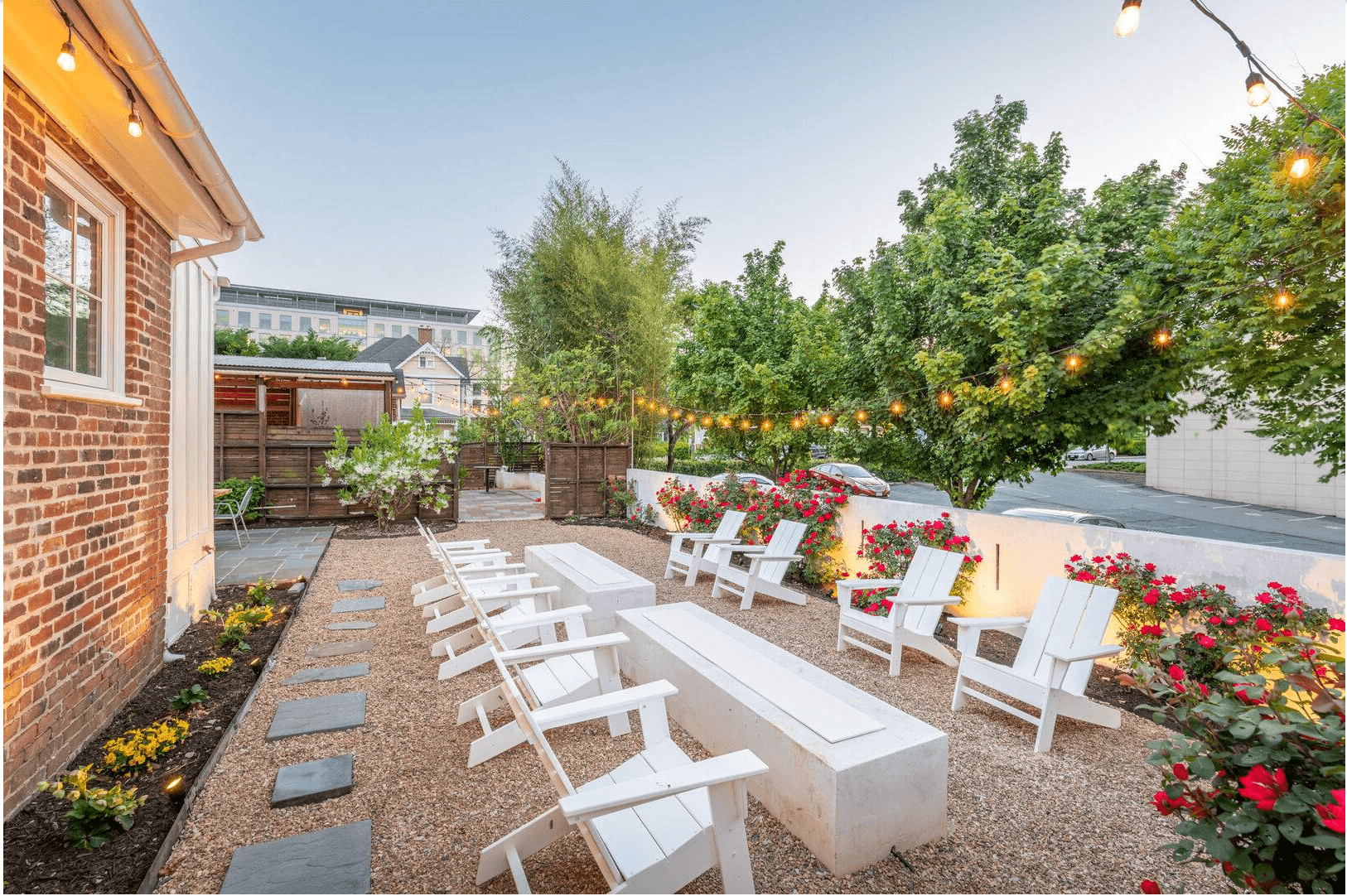 Outdoor patio with white chairs, picnic tables, string lights, and colorful flowers surrounded by trees and a brick building.