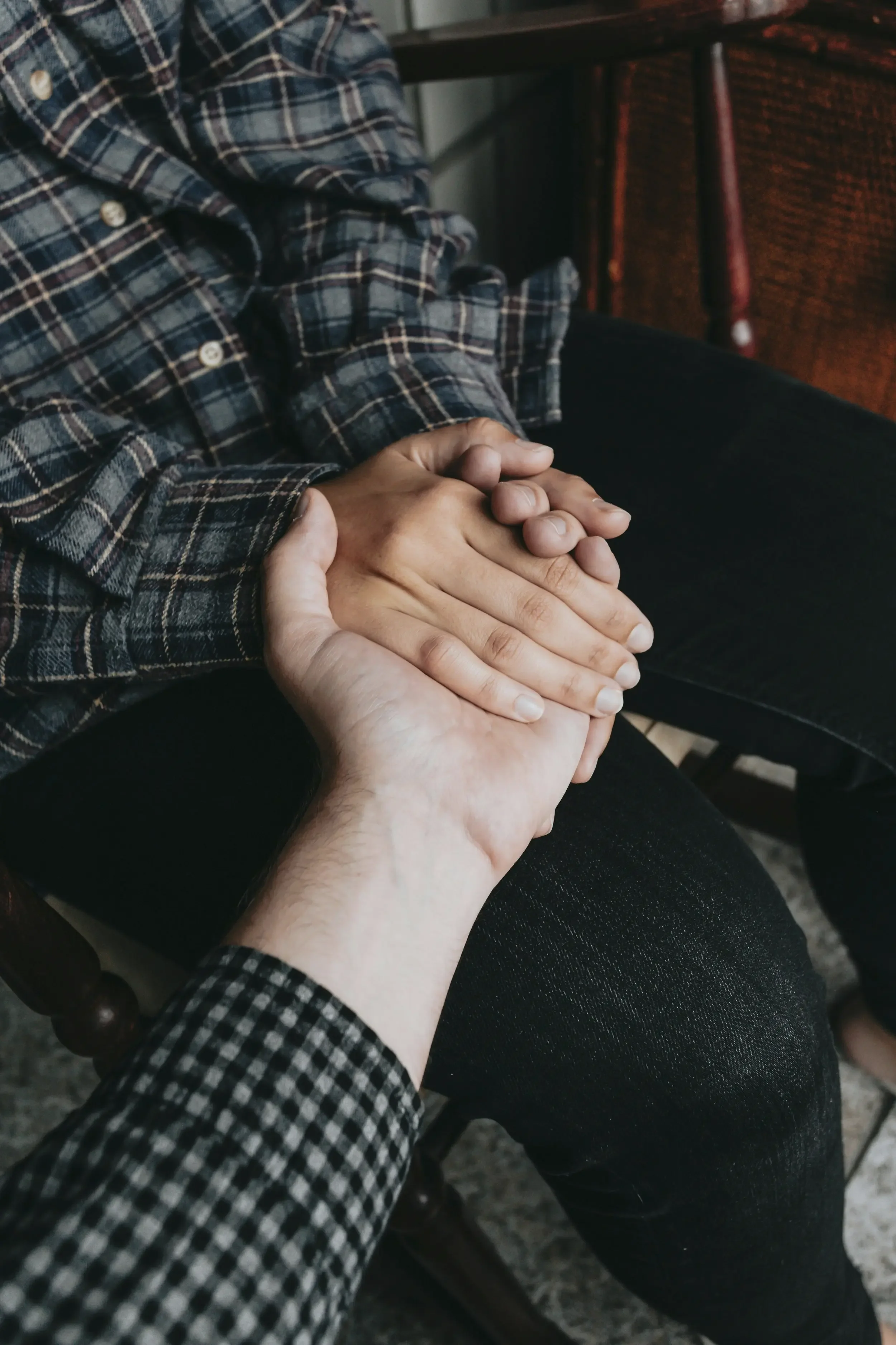 A person in a plaid shirt gently holding another person's hand. The person being held is wearing a similar plaid shirt and is seated on a wooden chair.