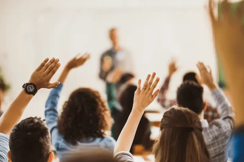 Group of students in a classroom raising hands to answer a question, with a teacher standing in the background