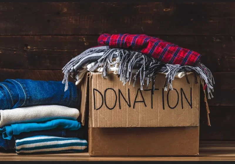 A cardboard box labeled 'Donation' with folded clothes and scarves stacked beside and draped over it on a wooden surface.