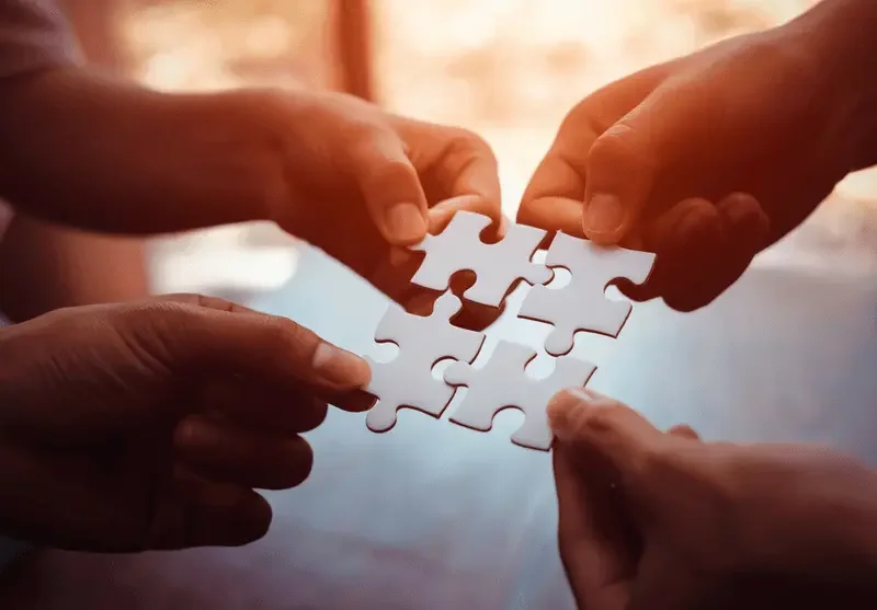 Four hands holding and connecting four white puzzle pieces together.