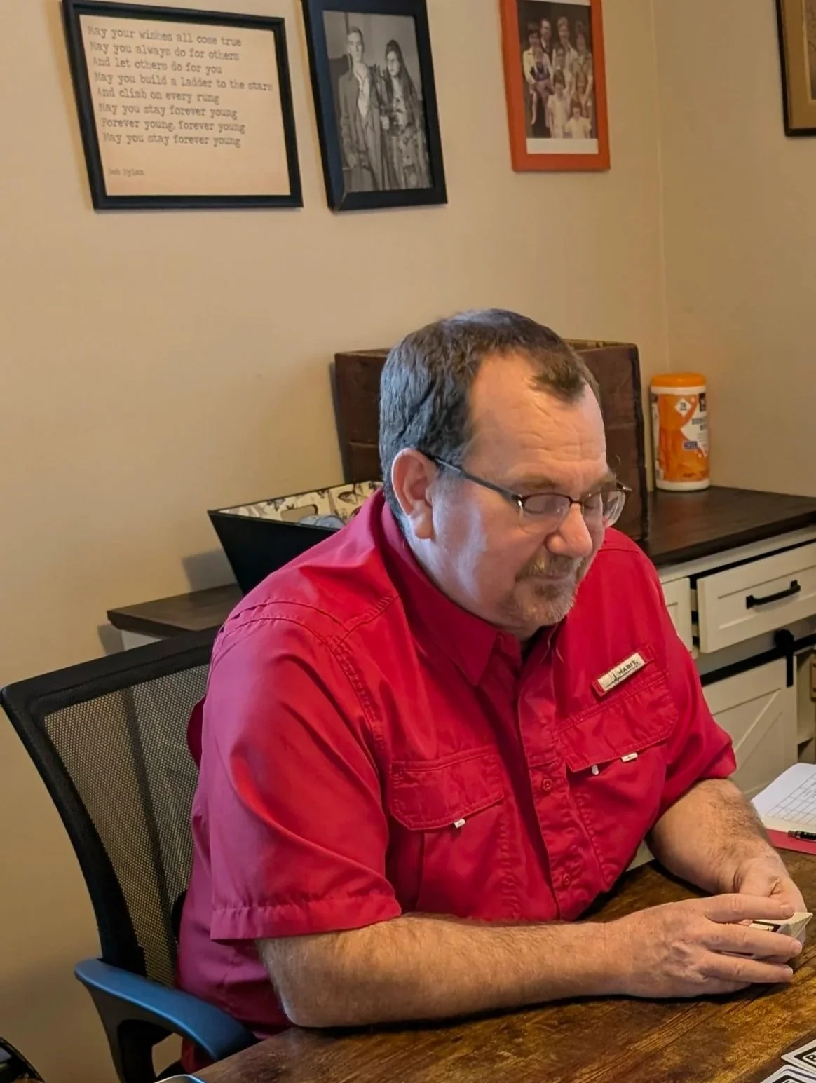 A man and a boy sitting at a wooden desk with educational materials and photographs on the wall in an office setting.