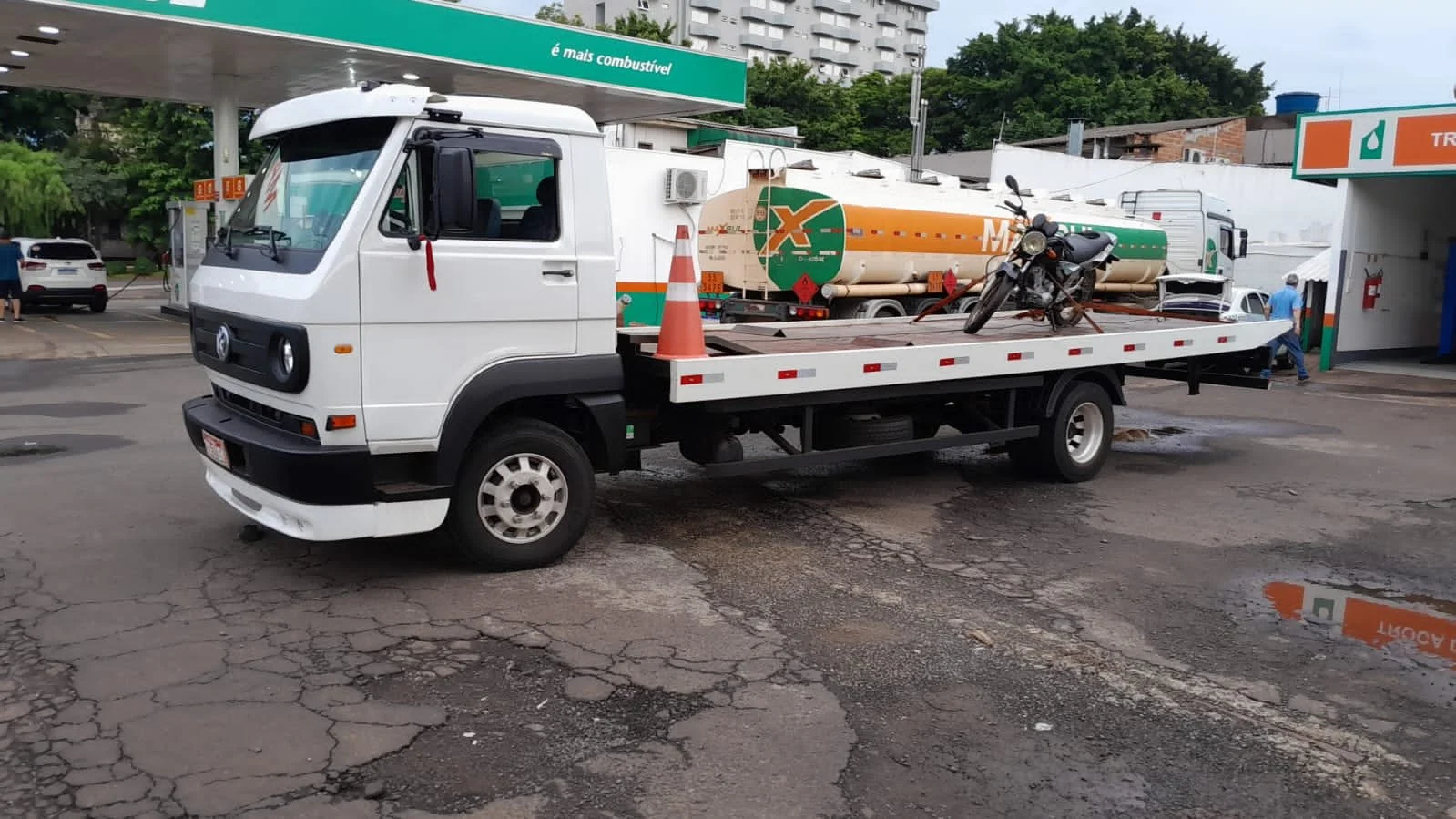 Caminhão da Am Guinchos, com uma moto na plataforma do caminhão e cones de trânsito, em um posto de gasolina
