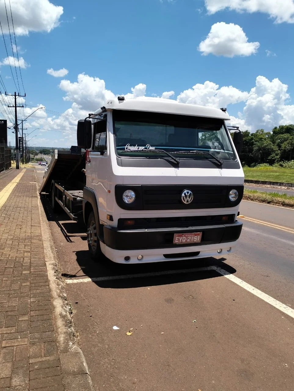 Caminhão Volkswagen branco estacionado na rua sob céu com nuvens brancas e céu azul, com árvores ao fundo.