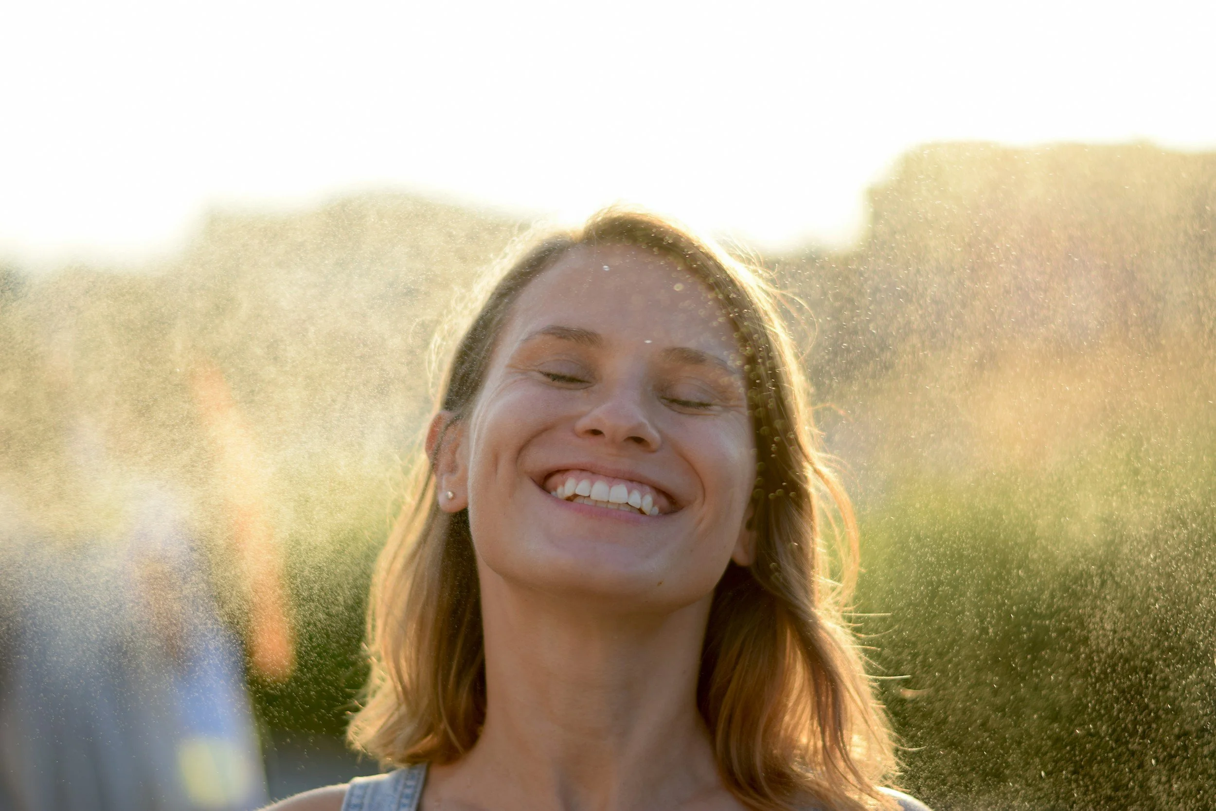 A white woman with eyes closed and smiling wide with a slight mist of water and hazy light background, showing the kind of relief that is possible with online anxiety therapy for women with anxiety in Greenville, SC