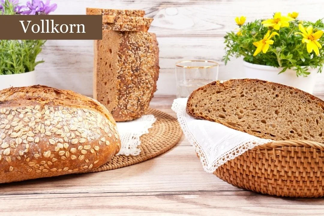 Verschiedene Brotsorten auf einem Tisch. Ein Haferflockenbrot liegt vorne, daneben ein dunkles Vollkornbrot, beide auf runden Tabletts. Zwei kleine Blumentöpfe mit gelben und lila Blumen stehen im Hintergrund. Der Text "Vollkorn" ist oben eingefügt.