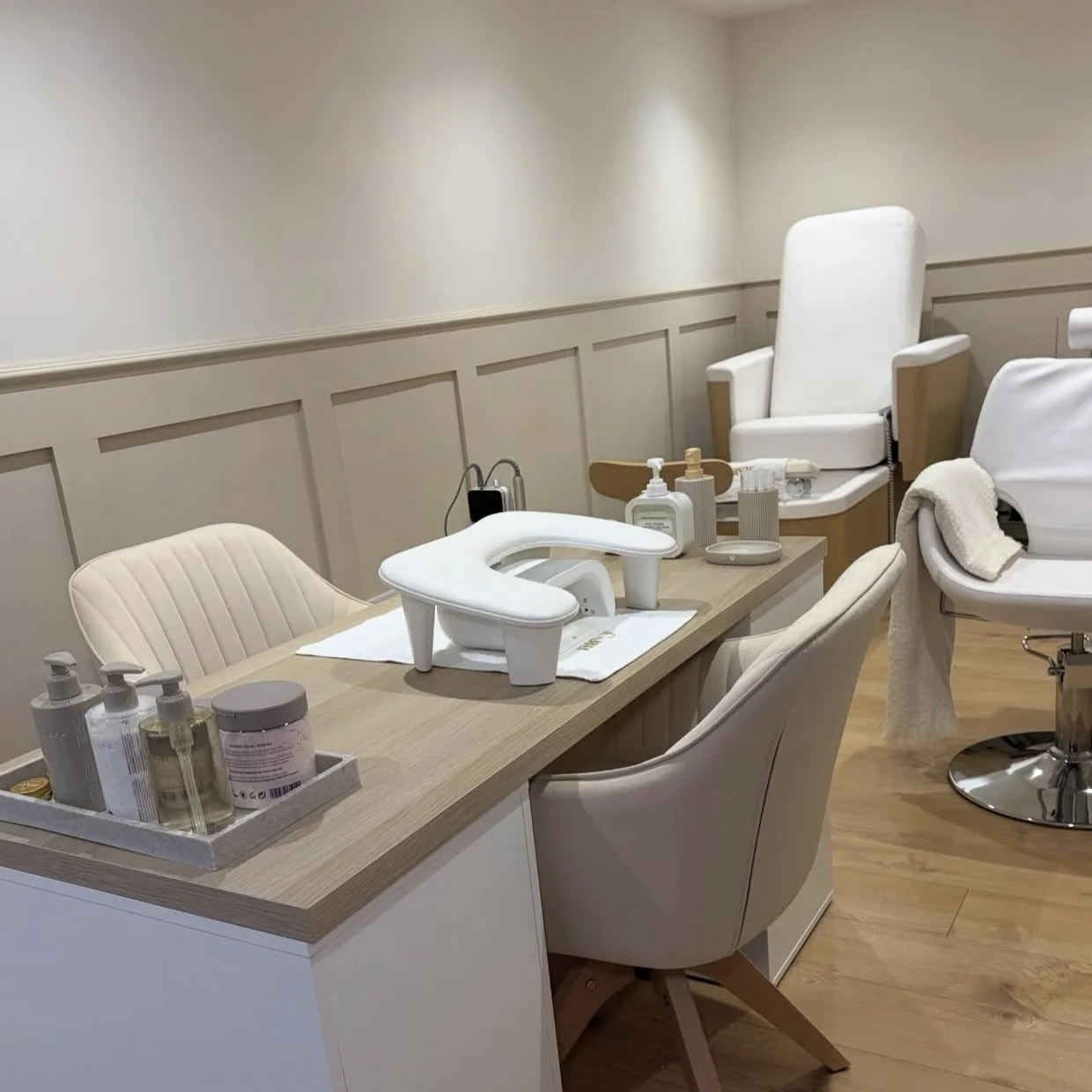 A medical or beauty treatment room with reclining chairs, a wooden table with skincare products and a machine, and a treatment bed with towels in the background.