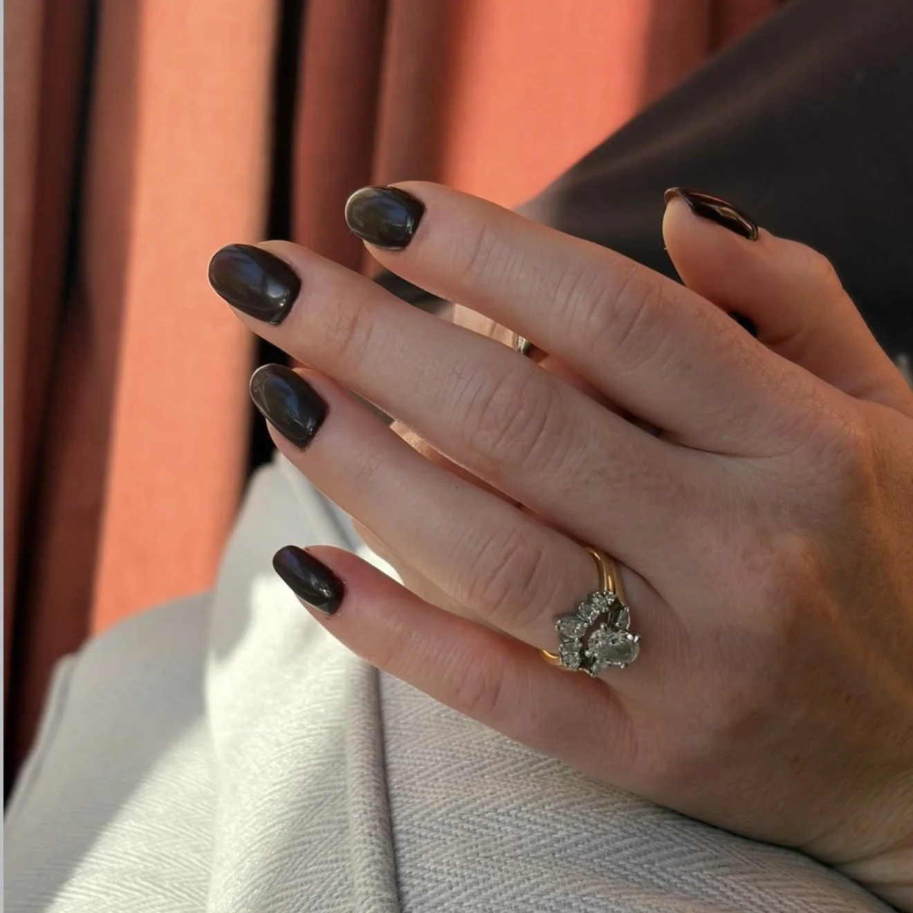 Close-up of a woman's hand showing a wedding ring with a cluster of diamonds and black painted nails, with a background of warm-colored curtains.