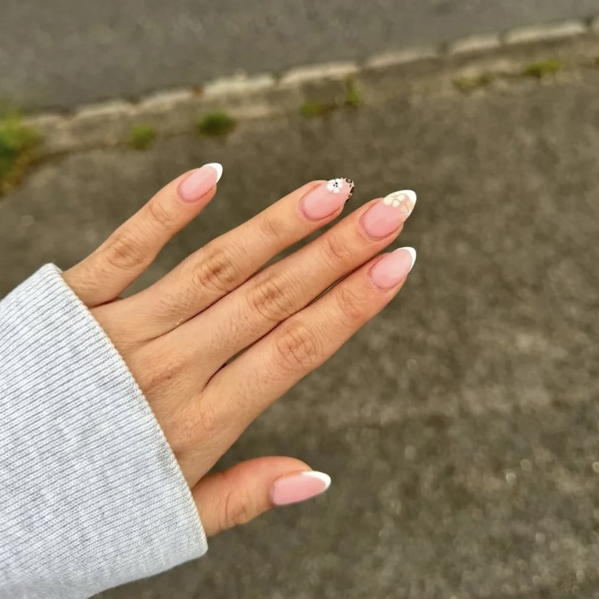 A hand with manicured nails featuring white tips and floral designs, held against a ground with grass and a sidewalk.