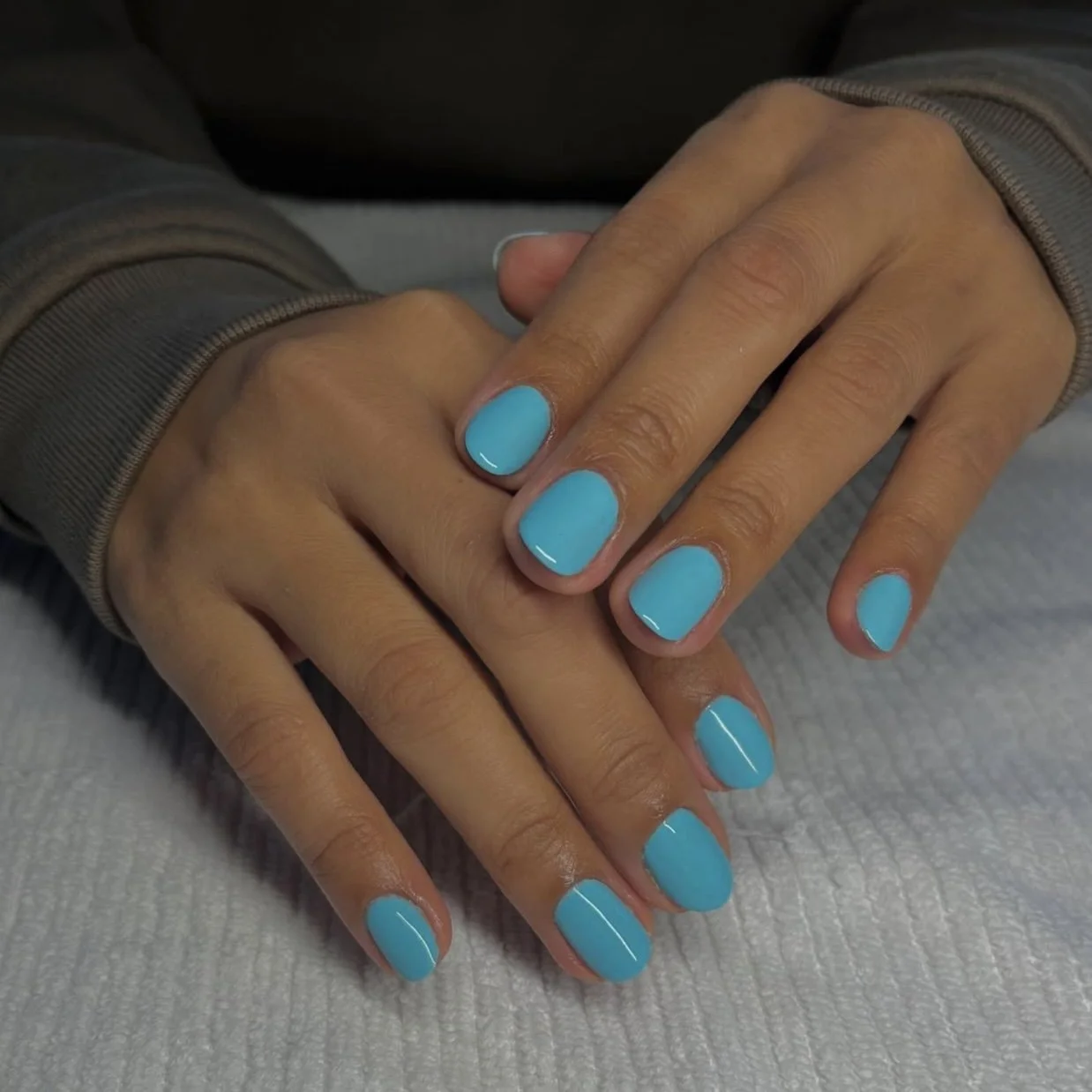 Close-up of hands with light blue painted nails, resting on a white textured surface, wearing a gray long-sleeve shirt.