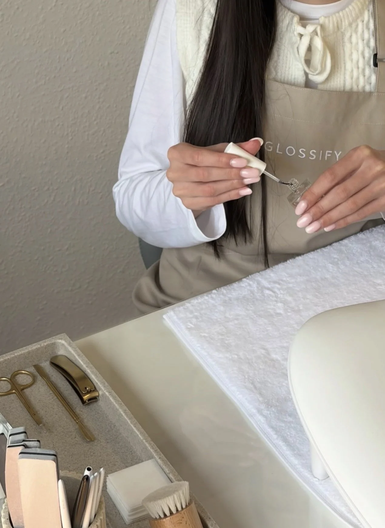A woman in a white long-sleeve shirt and a beige apron stands at a white counter, preparing a cosmetic or skincare product. The apron has the brand name 'GLOSSIFY' printed on it. On the counter, there are various beauty tools, including scissors, twe