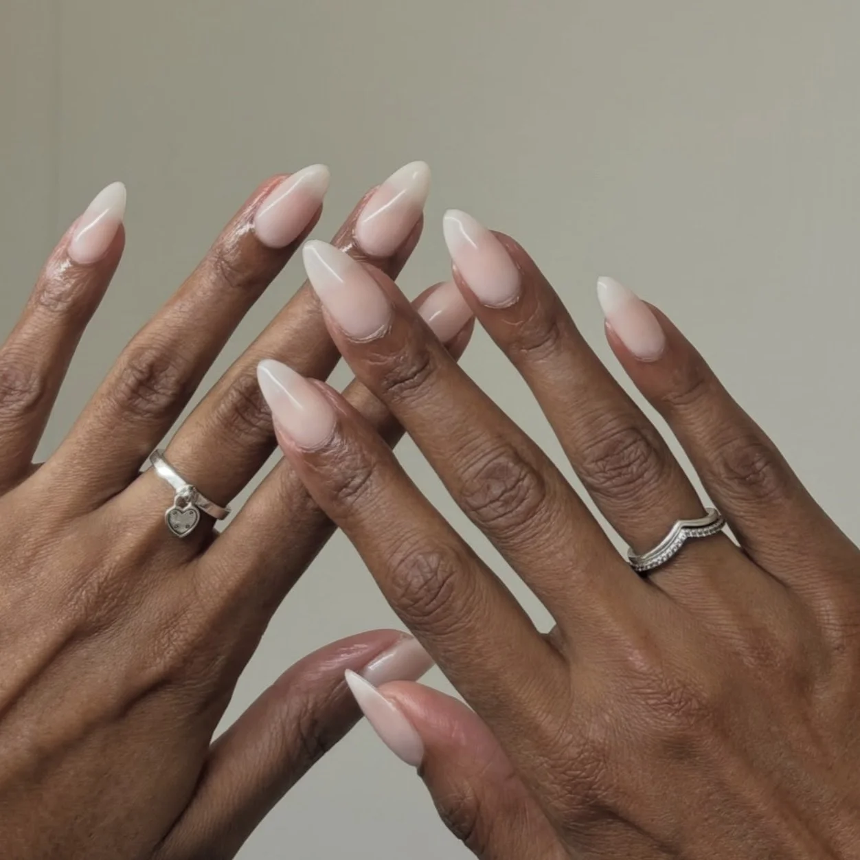 Two hands with well-manicured, nude-colored, almond-shaped nails, fingers adorned with silver rings, some with heart-shaped and engraved designs, against a plain background.