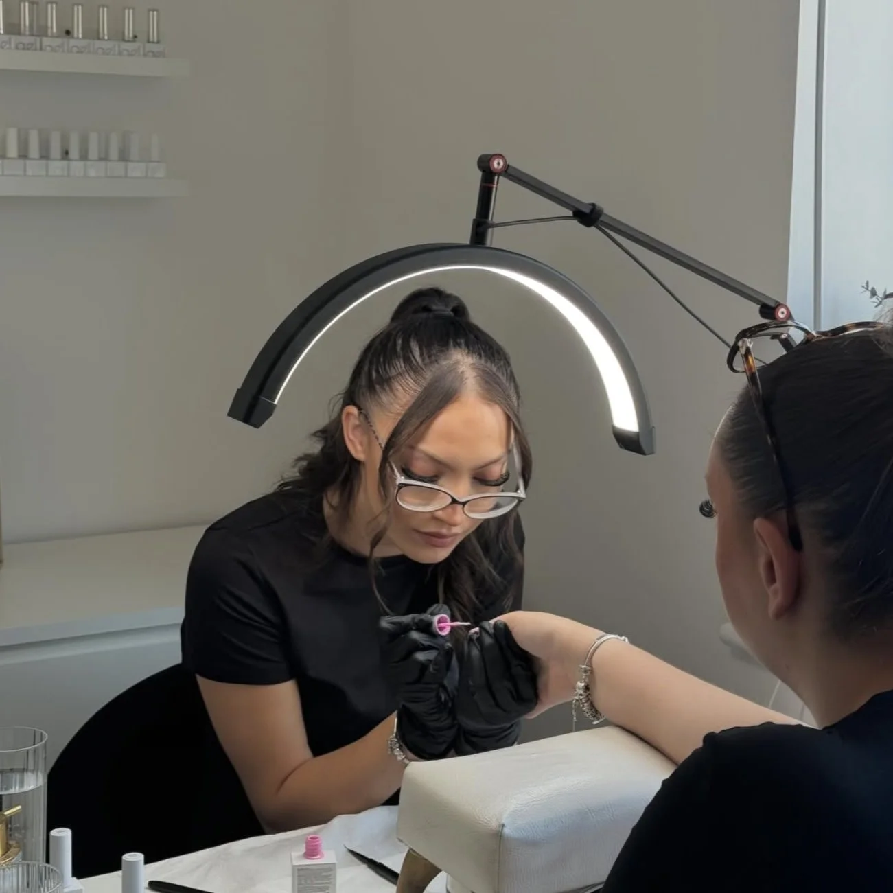 A woman with glasses getting a tattoo on her wrist from a tattoo artist in a salon, with tattoo supplies on the table and a large light above them.