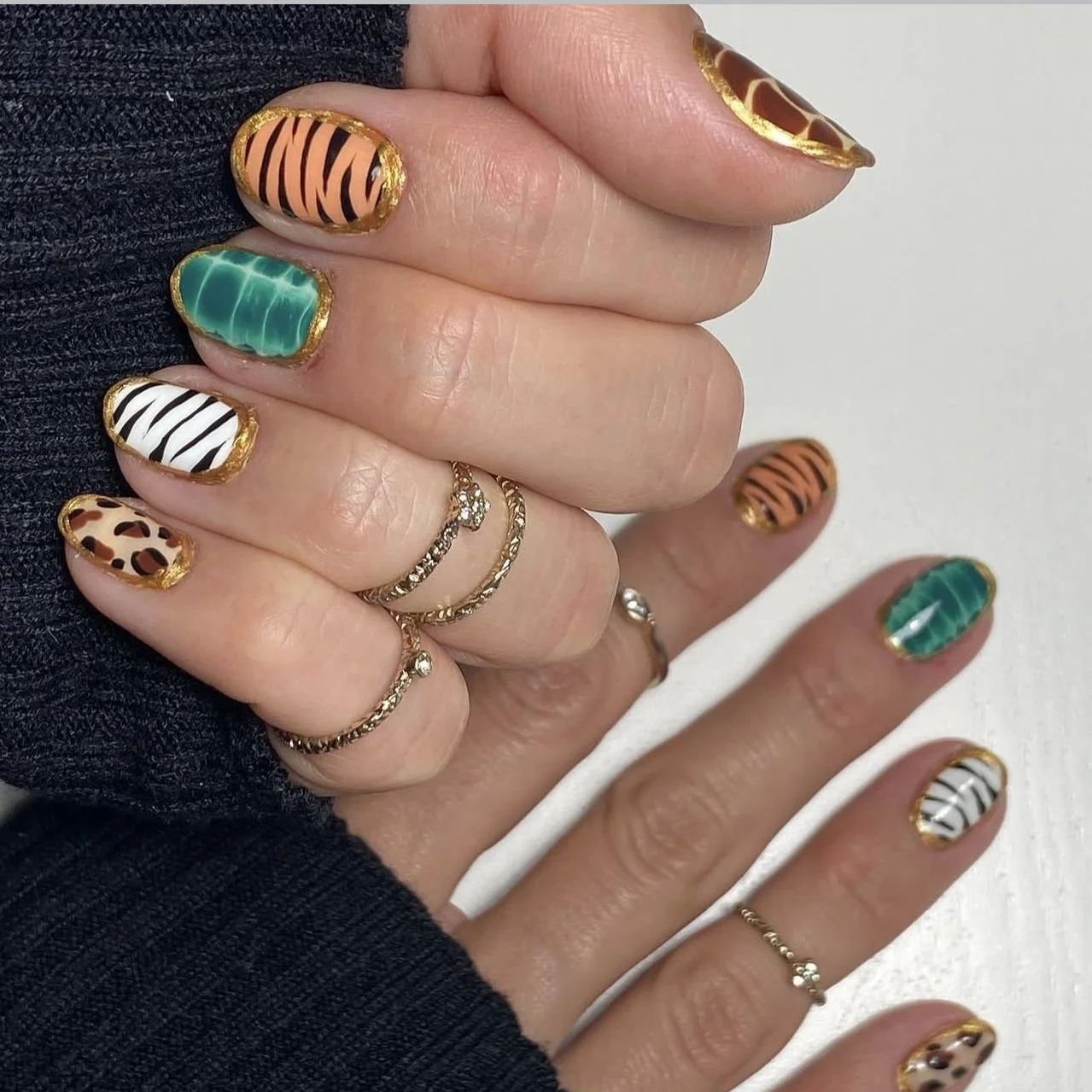 Hand with colorful animal print and zebra striped nail designs, some accented with gold borders, adorned with rings.
