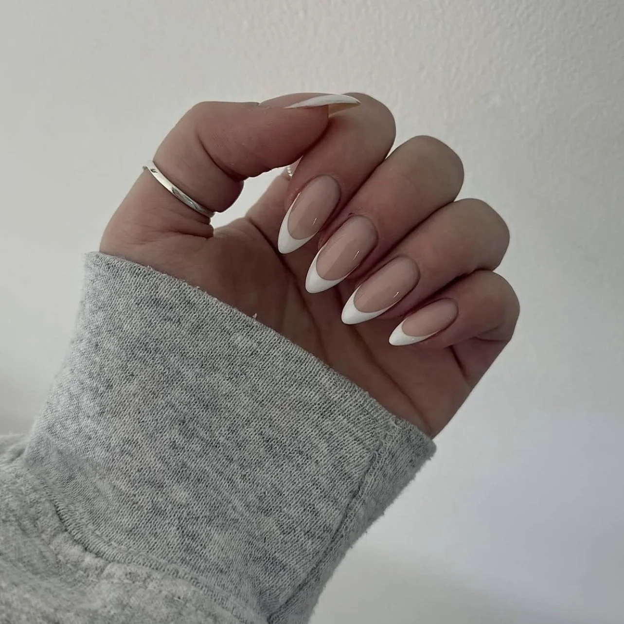 Close-up of a hand with manicured nails featuring a French tip design, wearing a silver ring and a grey long sleeve.