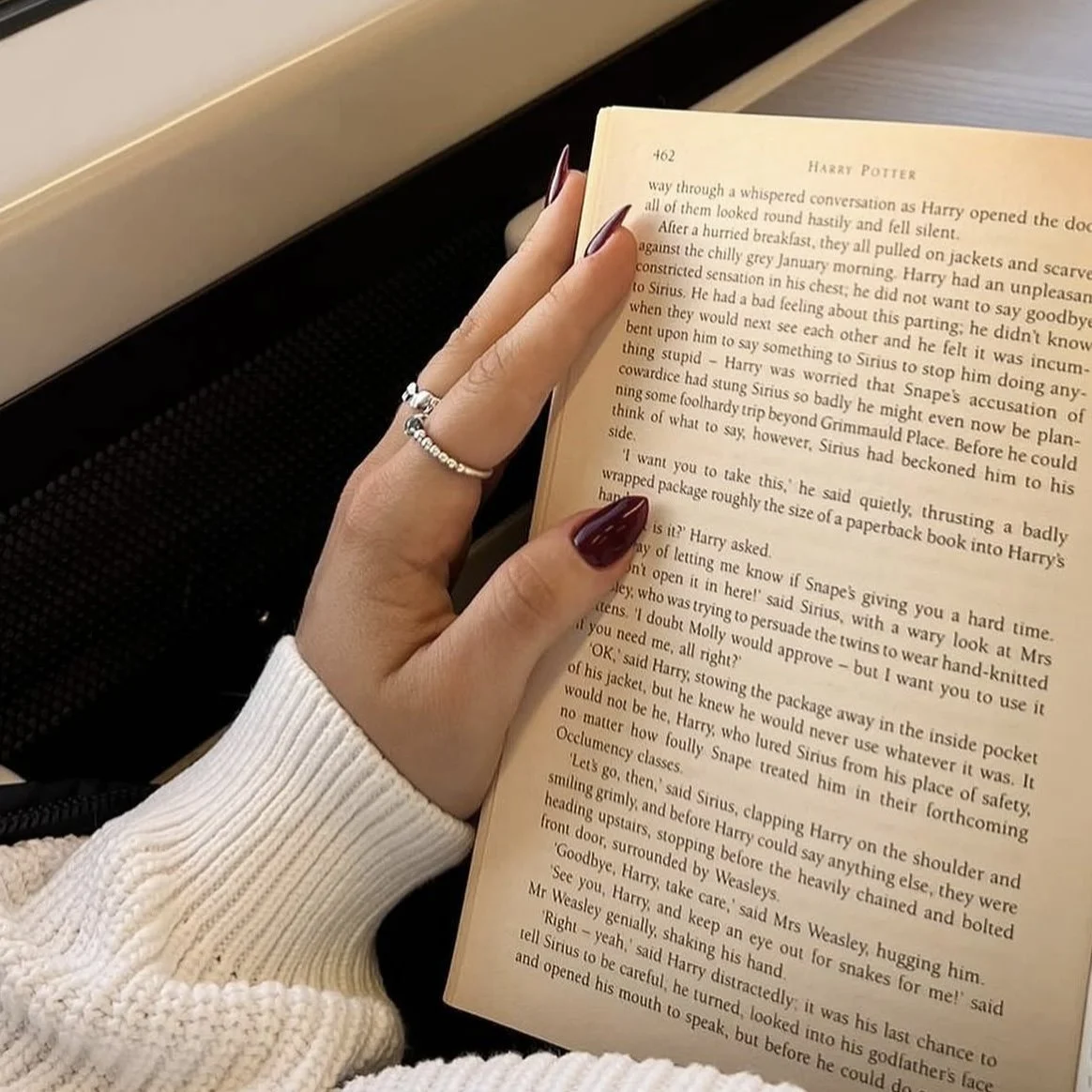 A person holding a paperback Harry Potter book, with their hand resting on the left page. The person is wearing a white ribbed sleeve and silver rings, with long, dark red painted nails.