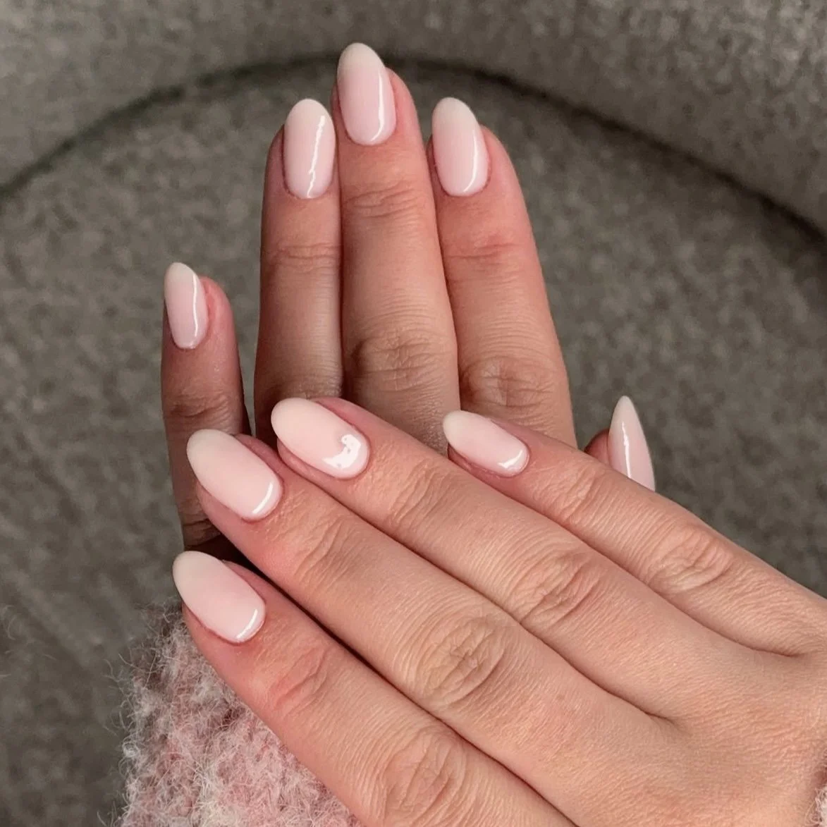Close-up of hands with neatly done nude-colored, oval-shaped, glossy manicured nails, overlapping each other, against a textured gray background.