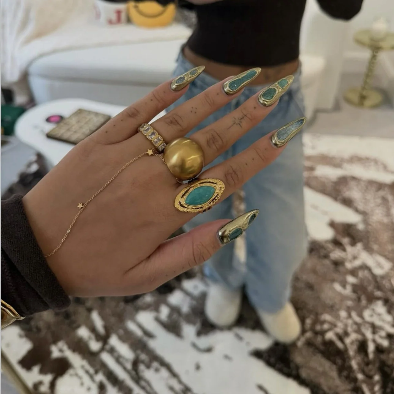 Person showing off multiple gold and turquoise jewelry rings, including a large gold ring, a turquoise and gold ring, a gold ring with clear stones, and a delicate gold bracelet with star charms. The person's nails are decorated with gold and turquoi