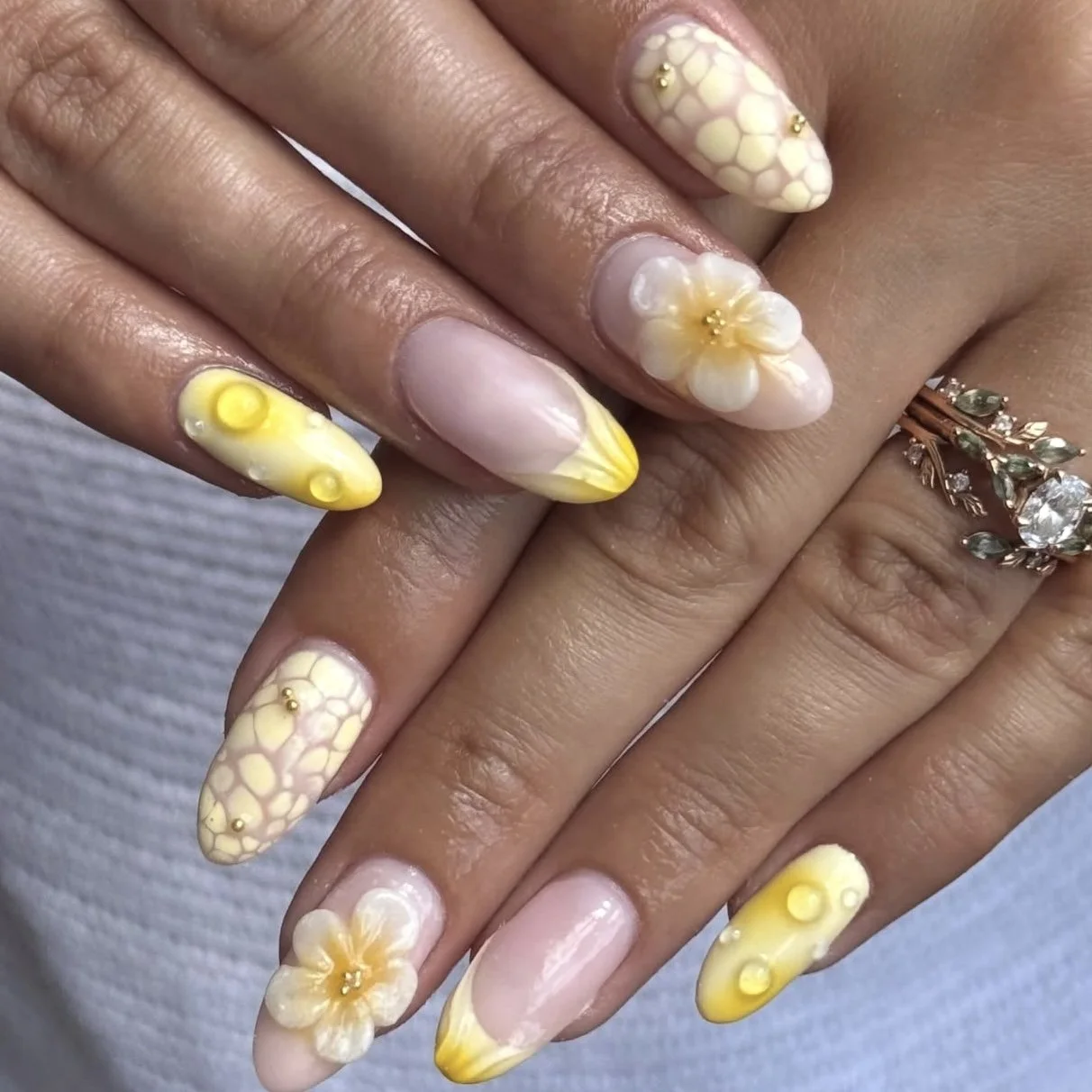 Close-up of hands with detailed yellow and white floral and polka dot nail art on long, almond-shaped nails, accessorized with gold and pearl embellishments.