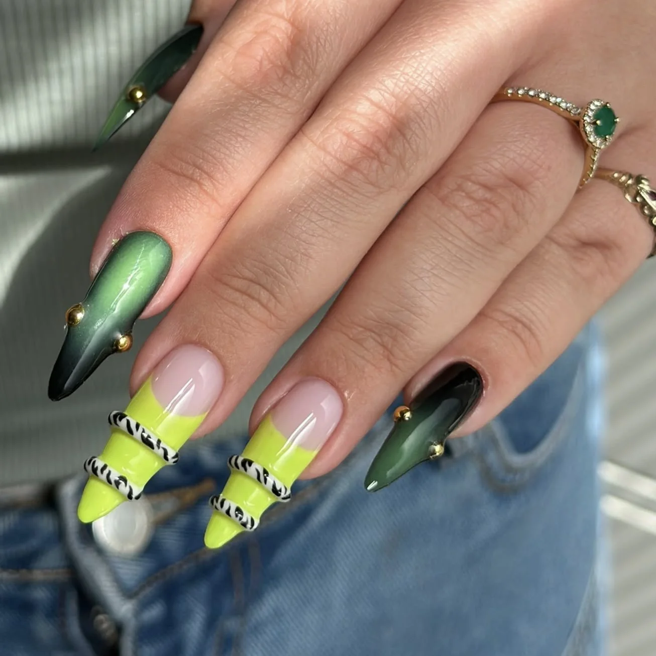 Hand with long, curved nails painted in black, green, and a neon yellow and black zebra pattern, wearing rings with green gemstones, and denim jeans visible in the background.