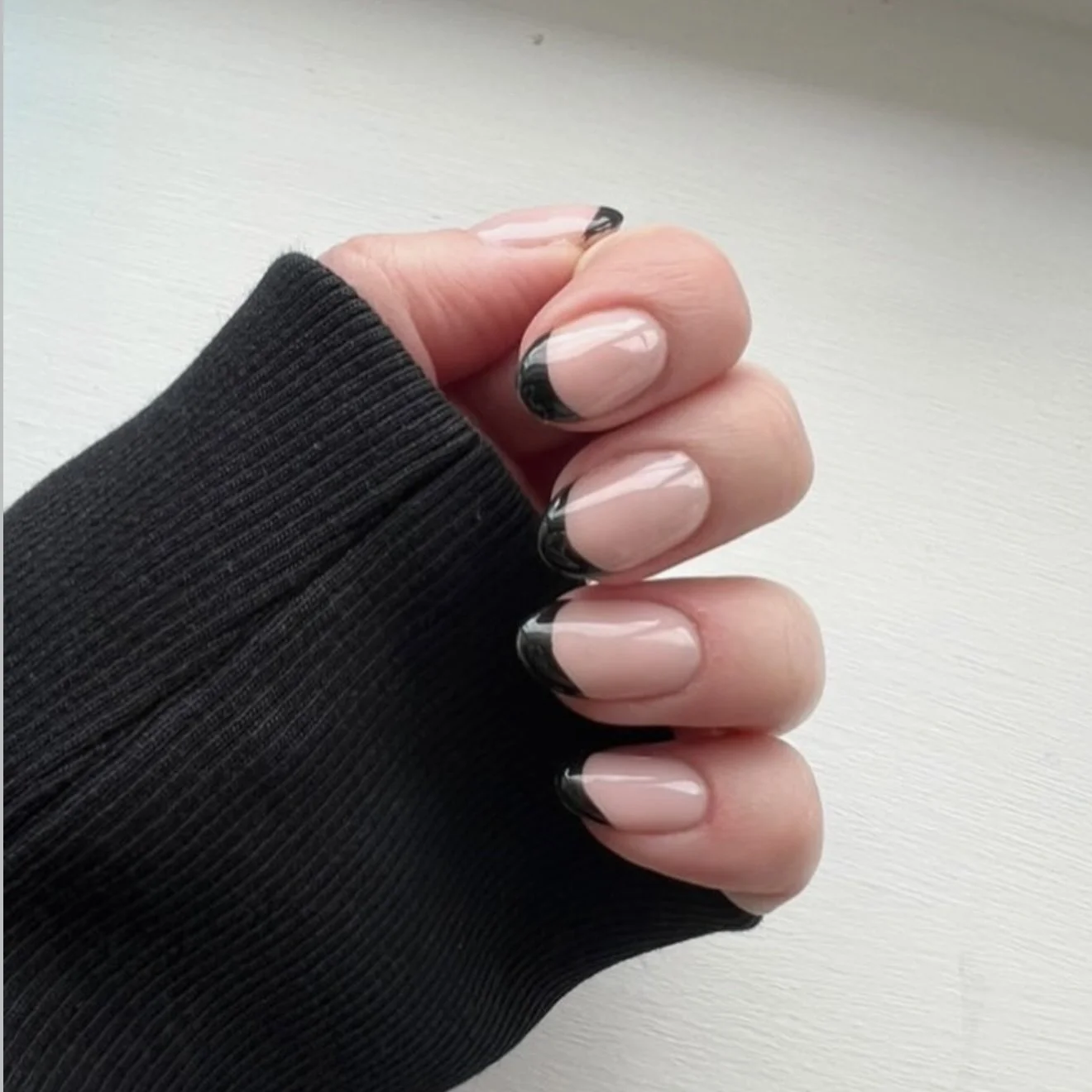 Close-up of a hand with black-framed nails in a French manicure style, wearing a black knitted sleeve with ribbing.