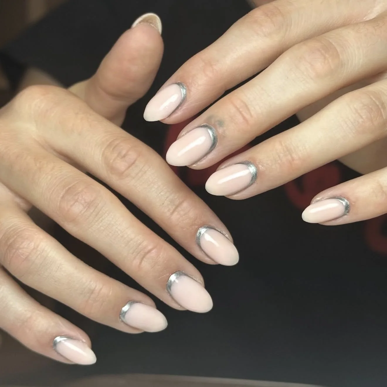 Close-up of hands with manicured nails that are painted in a light pink or nude shade with metallic silver tips, resting on a dark surface.