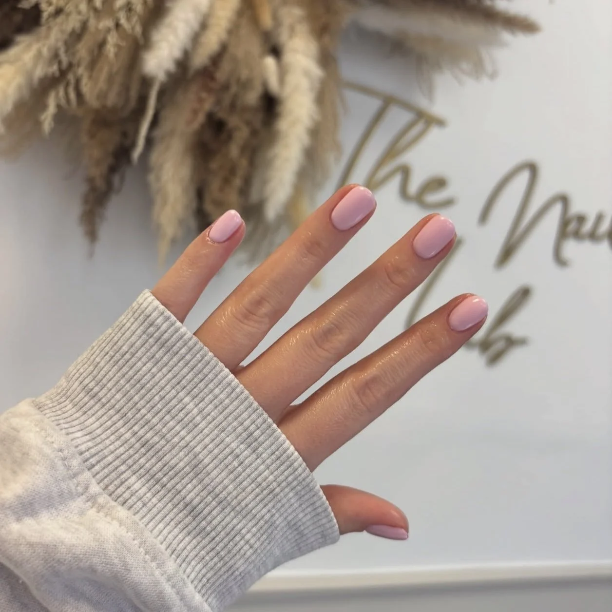A hand with neatly manicured nails painted in soft pink. The person is wearing a light gray, ribbed sleeve. In the background, there is a blurred wall with some dried pampas grass and a cursive sign that says 'The Mau'.
