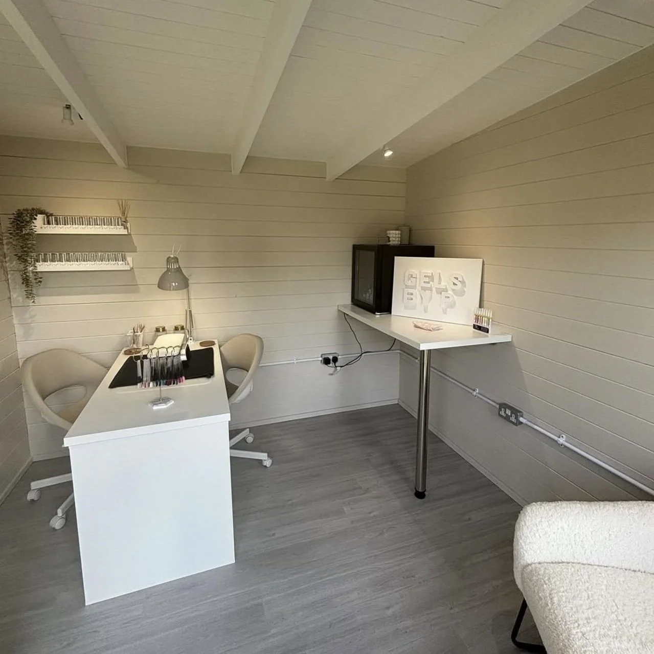 A small room with light-colored wooden walls and ceiling. There is a white desk with ergonomic chairs, nail polish bottles, and a desk lamp. On the wall, there is a white shelf with greenery and a small keyboard decor. On the right, a white tabletop 