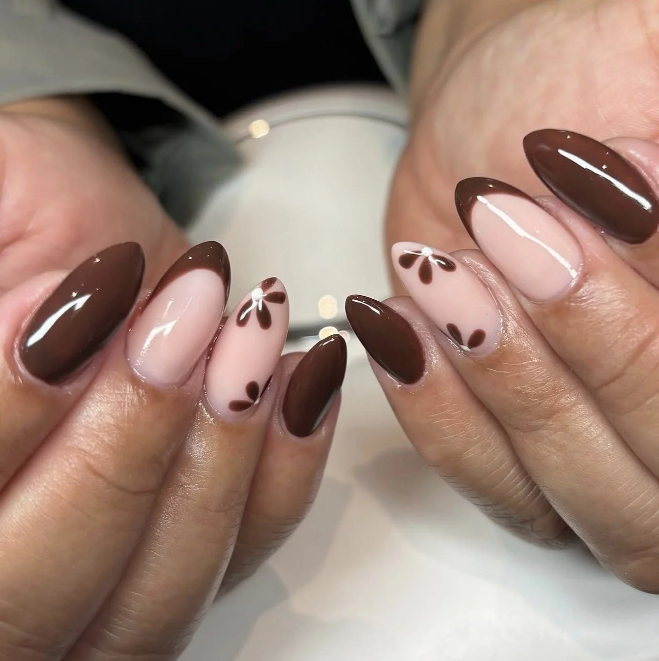 Close-up of hands with manicured nails featuring brown and nude colors, some nails with floral designs, held near a round mirror.