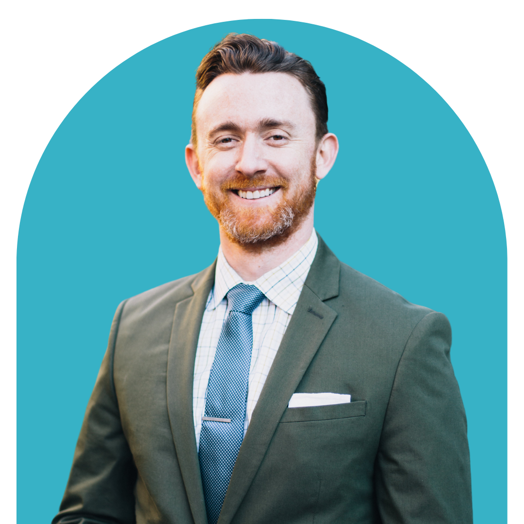 A smiling man with a beard and brown hair wearing a gray suit, light blue shirt, and blue tie, posing against a blue background.