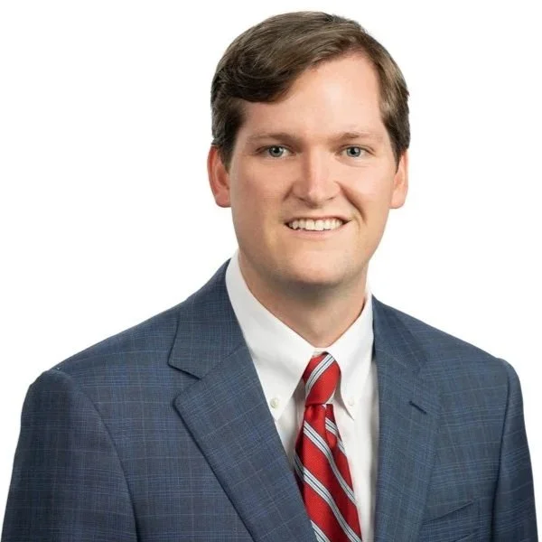 A professional portrait of a man with short brown hair, wearing a dark blue plaid suit, white shirt, and a red striped tie, smiling at the camera.