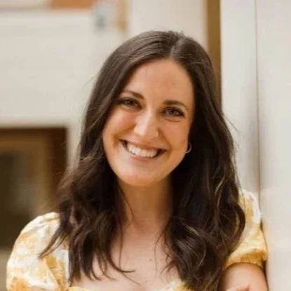 A woman with long, wavy brown hair and a wide smile, wearing a yellow floral top, standing indoors.