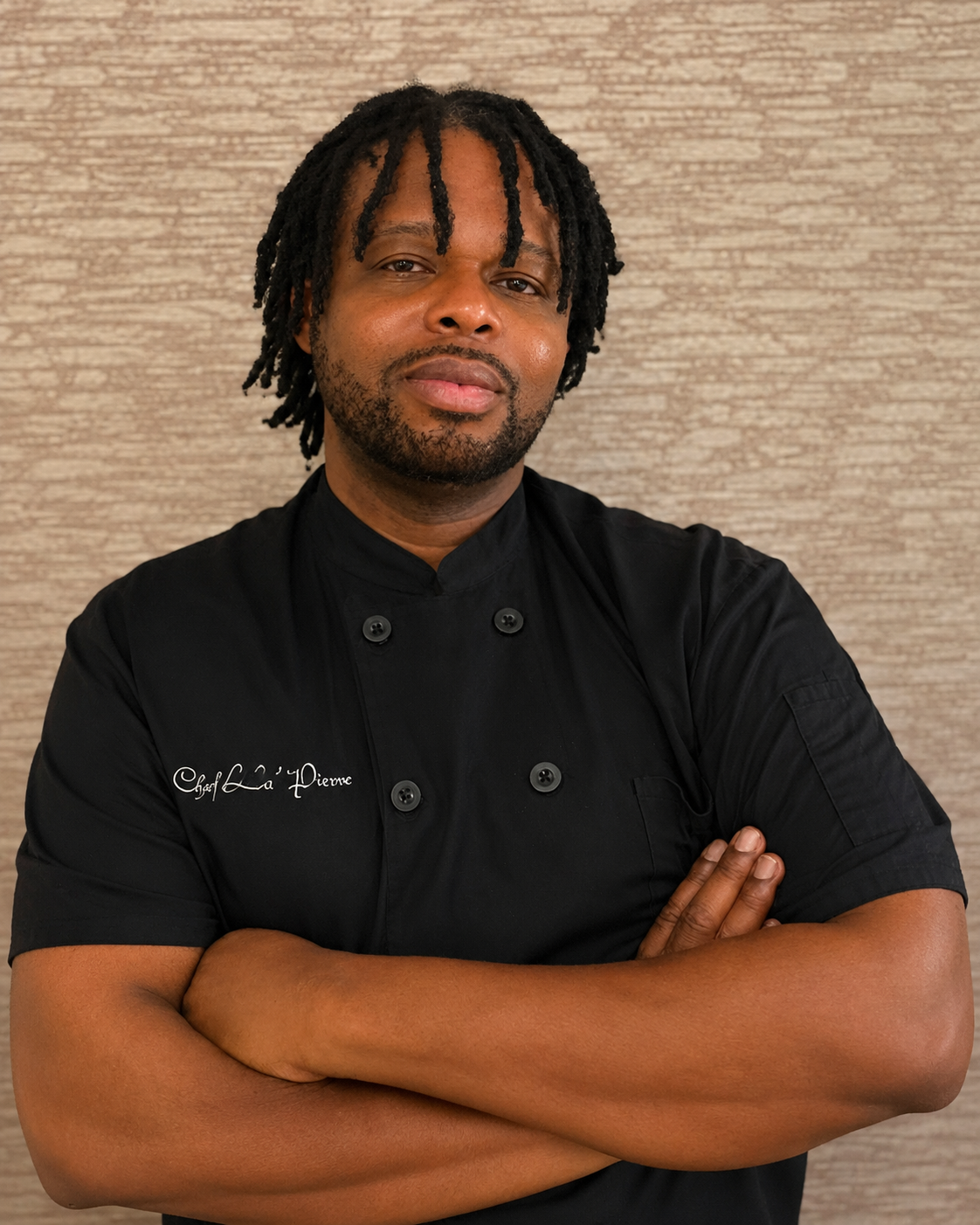Male chef in black uniform with crossed arms, standing against beige textured wall.