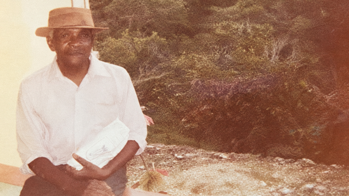 A man wearing a white shirt and a hat, standing outdoors in front of green foliage, holding a folder or paper.