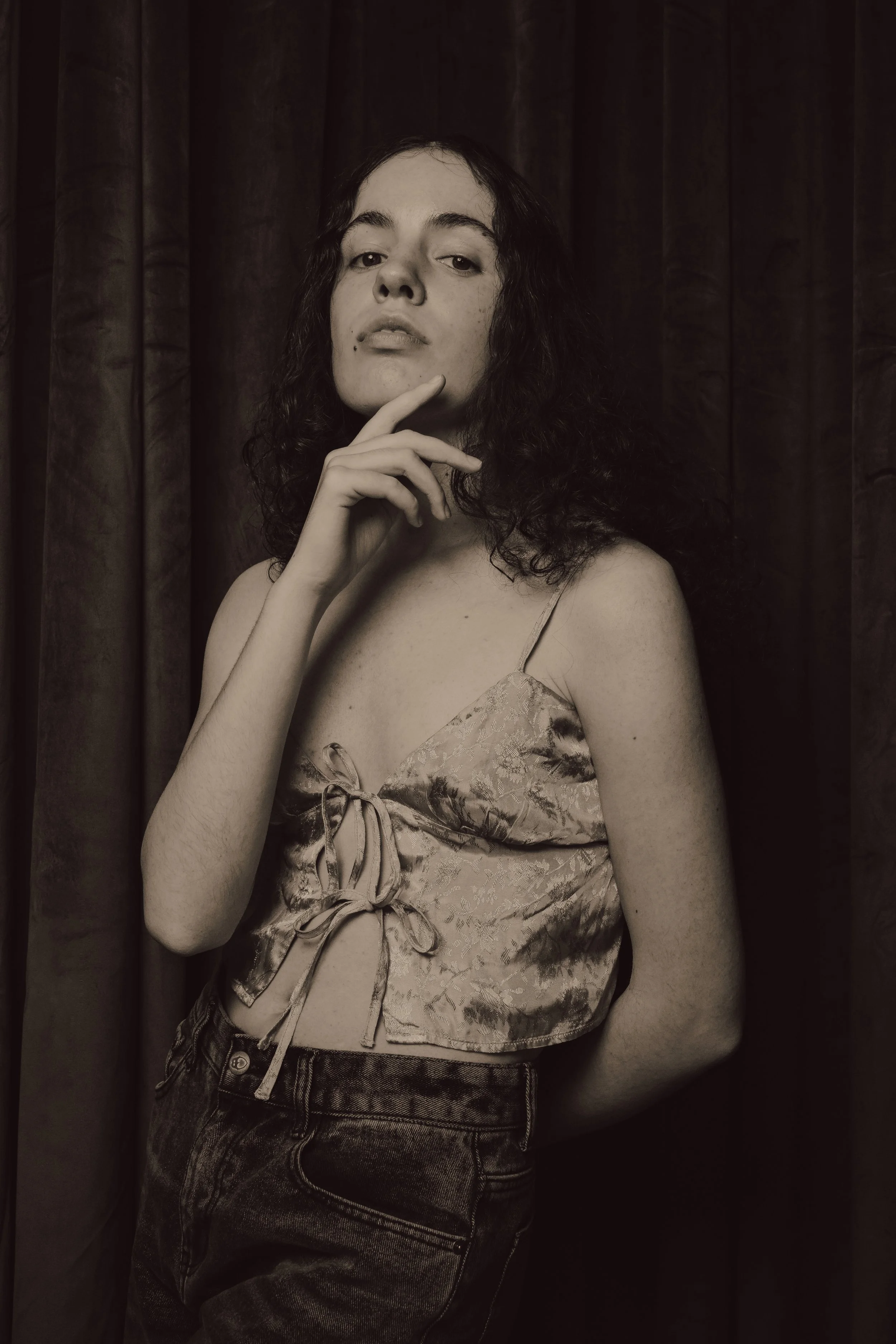 A woman with curly hair posing in front of dark curtains. She is wearing a patterned satin top and denim jeans, with her hand resting on her chin.
