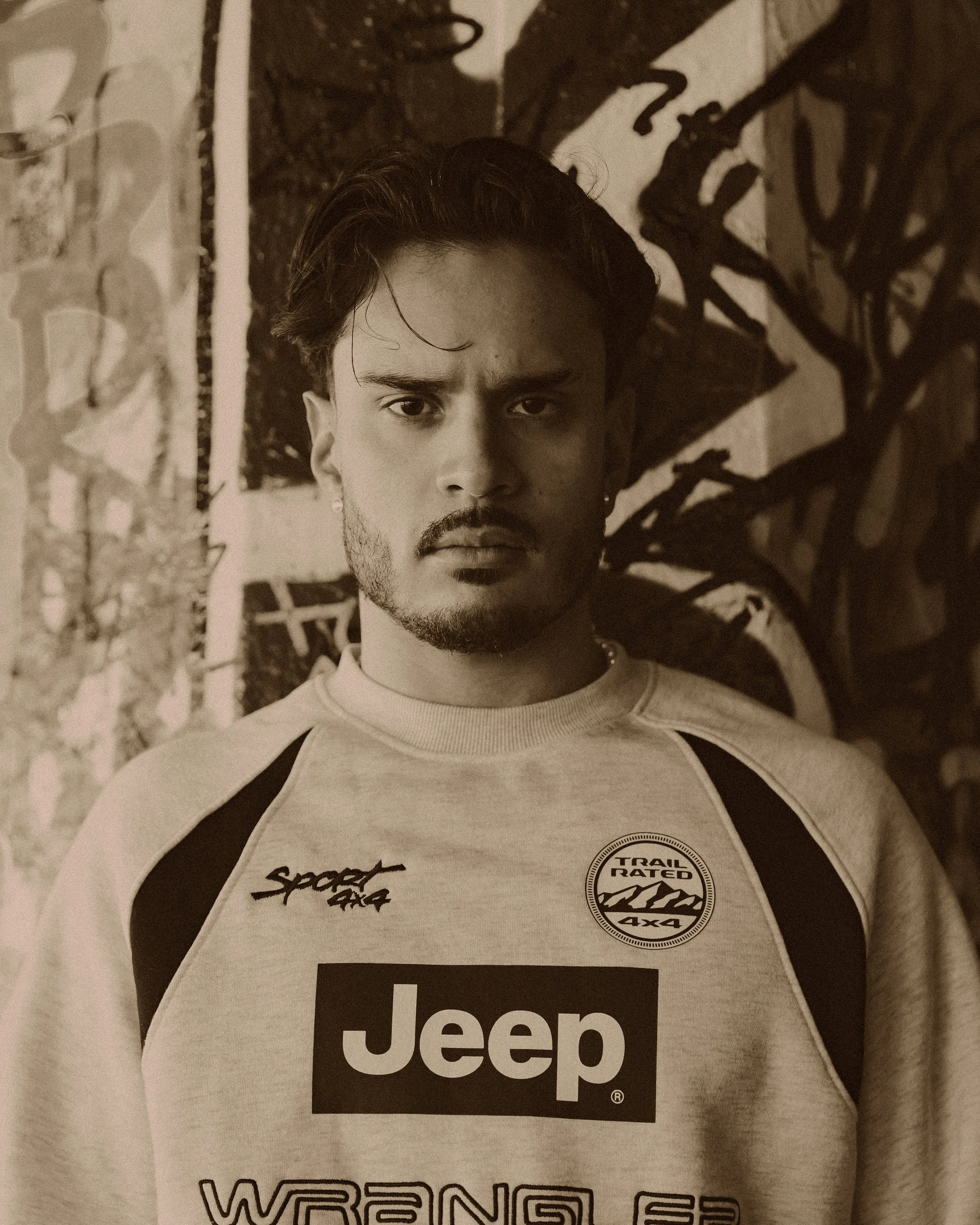 Young man with dark hair and facial hair, wearing a light-colored shirt with black sleeves, sitting in front of graffiti-style wall art.