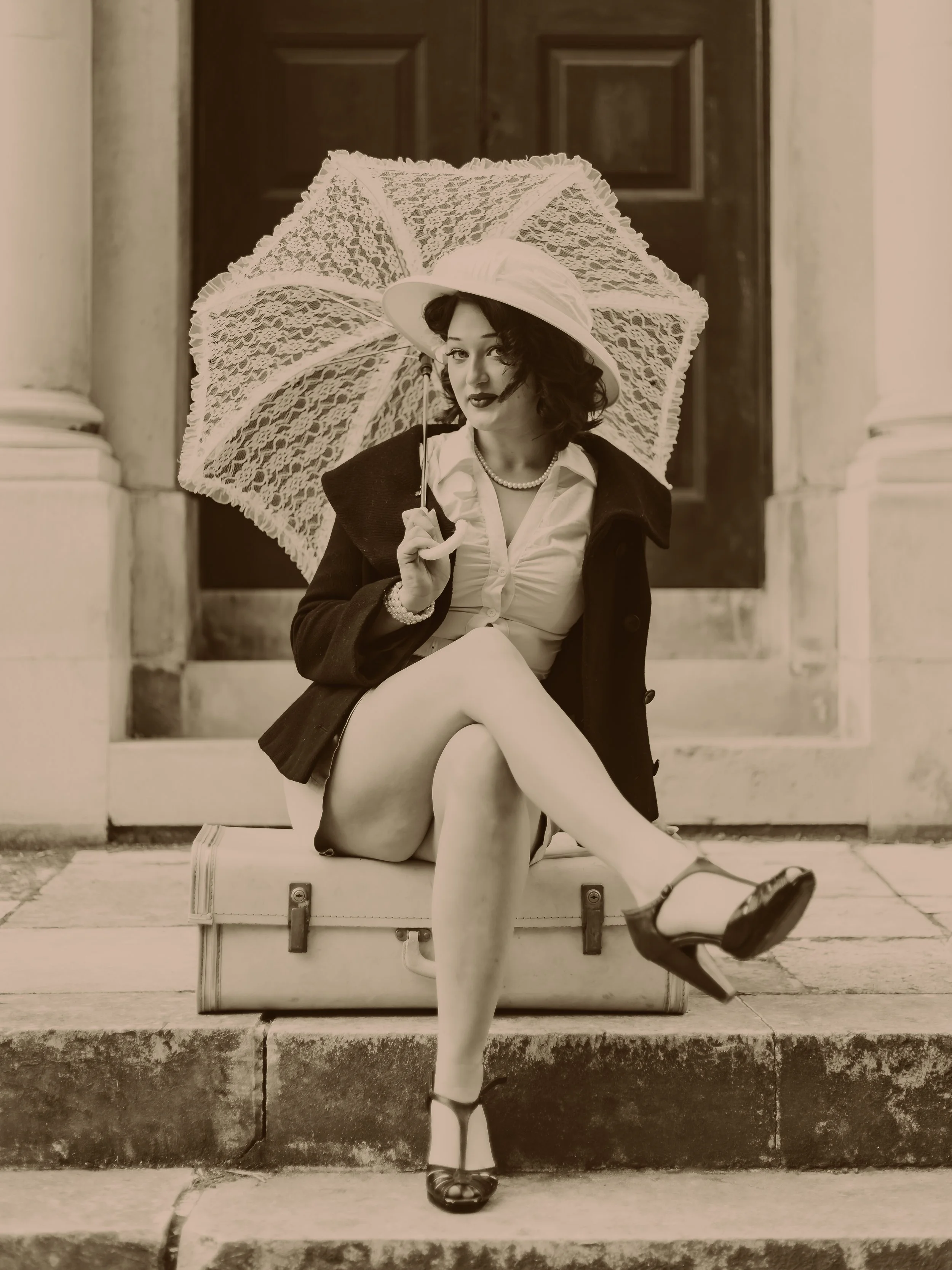 A woman dressed in vintage clothing sitting on a suitcase on steps, holding a lace umbrella, with a large hat, pearls, and high heels, in front of a building with columns and a door.