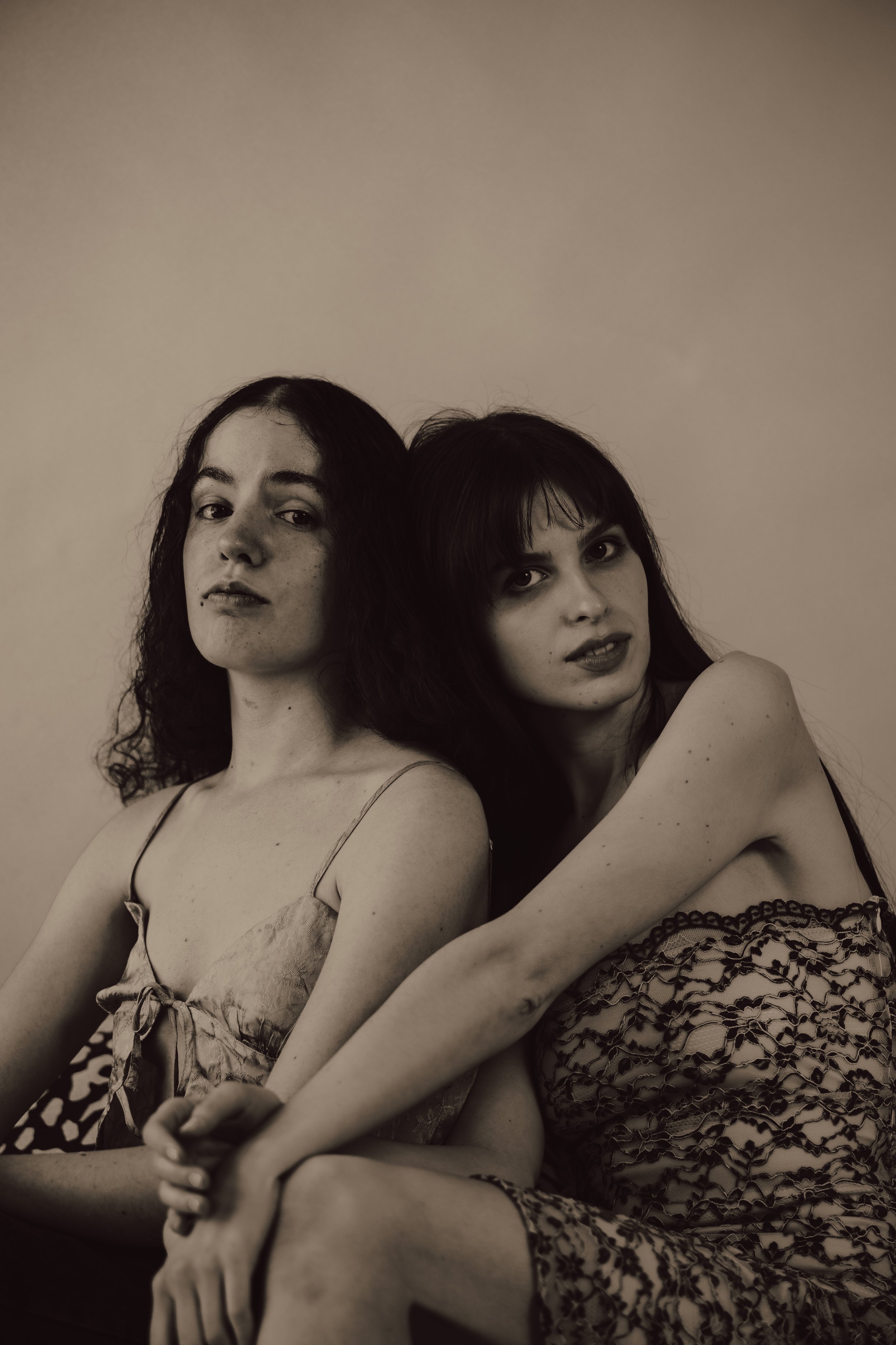 Two women sitting close together, one with curly dark hair and the other with straight dark hair and bangs, both wearing dresses with thin straps, against a plain background.