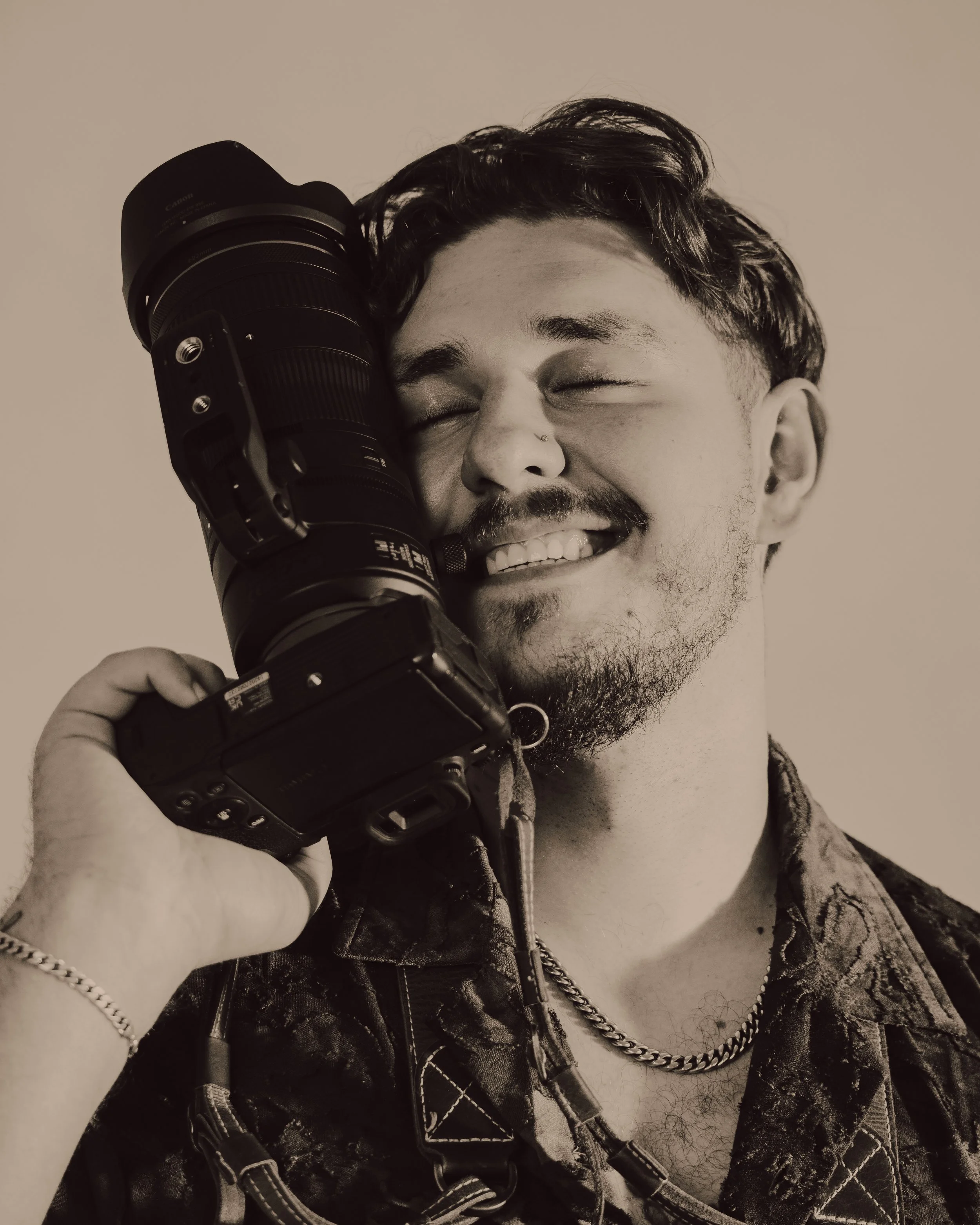 Black and white photo of a smiling person holding a professional camera lens to their face, with eyes closed, wearing a denim jacket, a chain necklace, and a beaded bracelet.