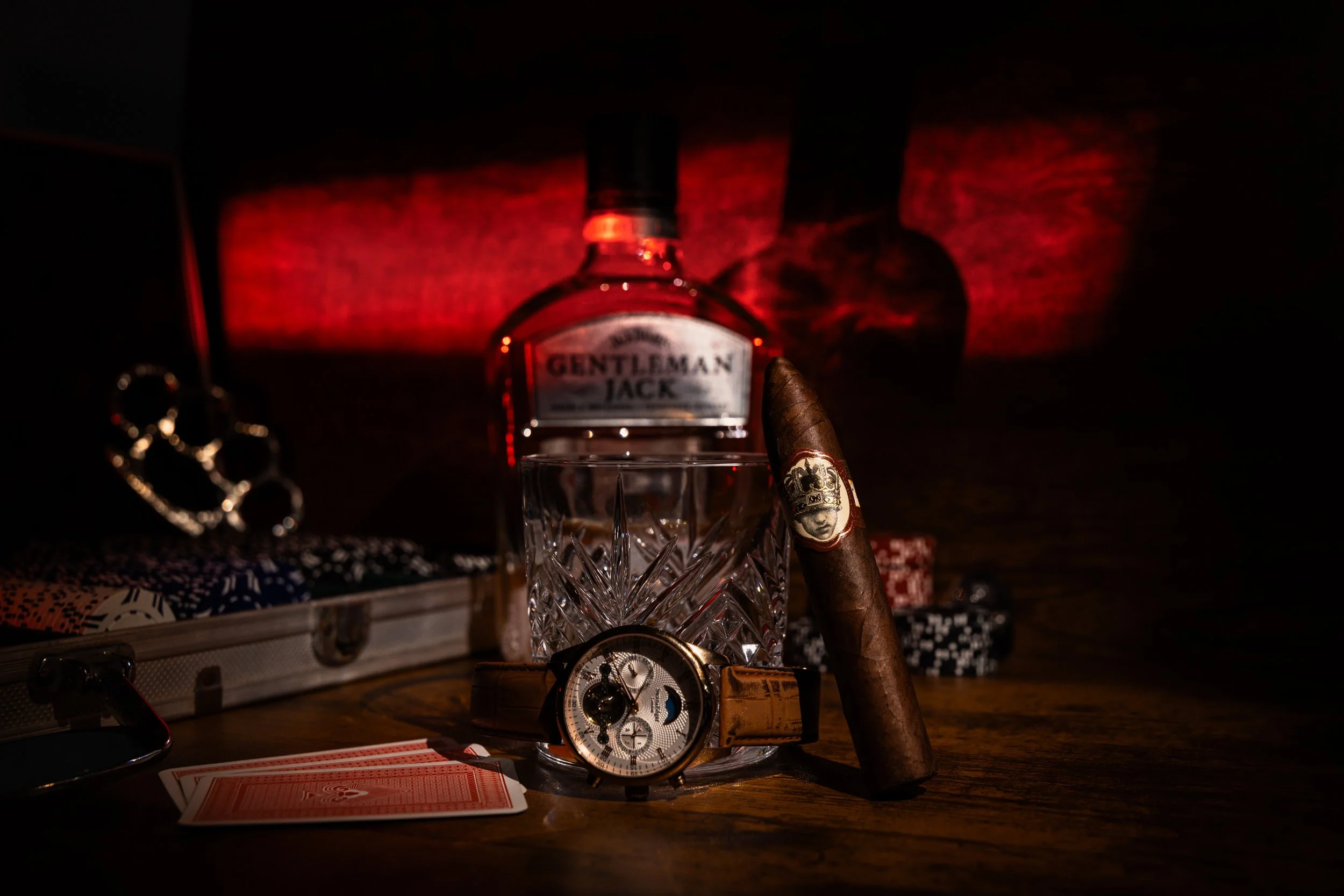 Cigar and a cut-crystal tumbler on a card table with a pocket watch, playing cards and chips, lit deep red