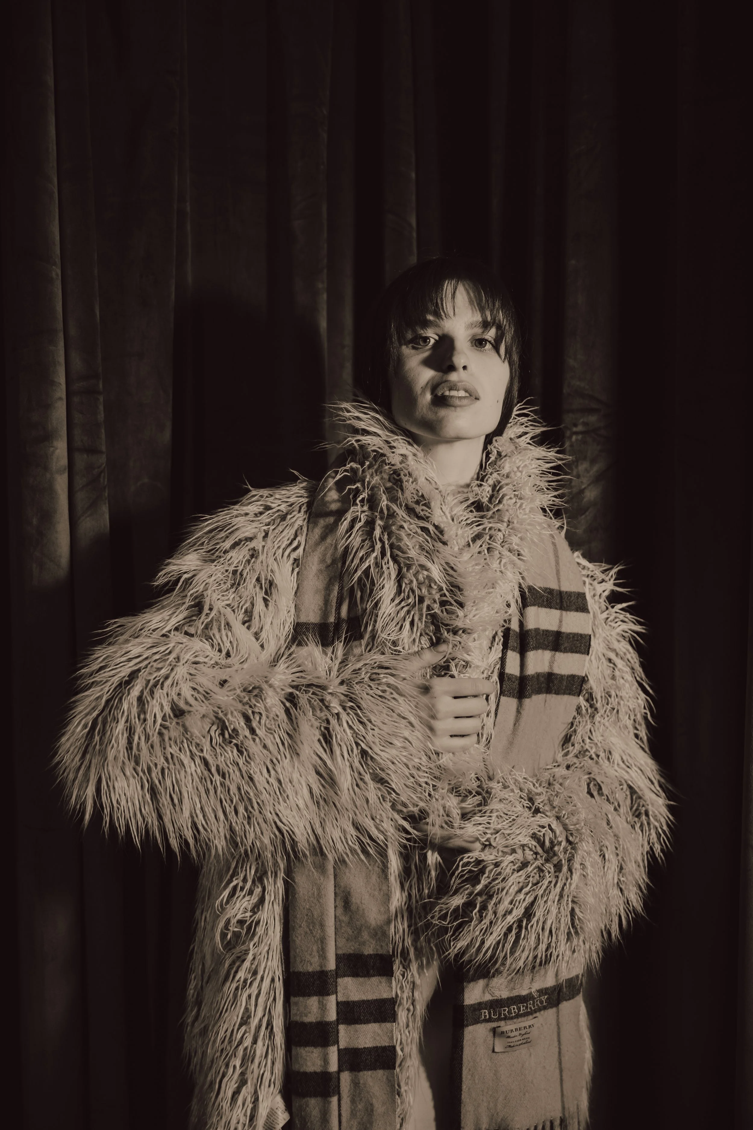 Black and white photo of a woman with short dark hair wearing a thick furry coat and a striped scarf, posing in front of dark curtains.