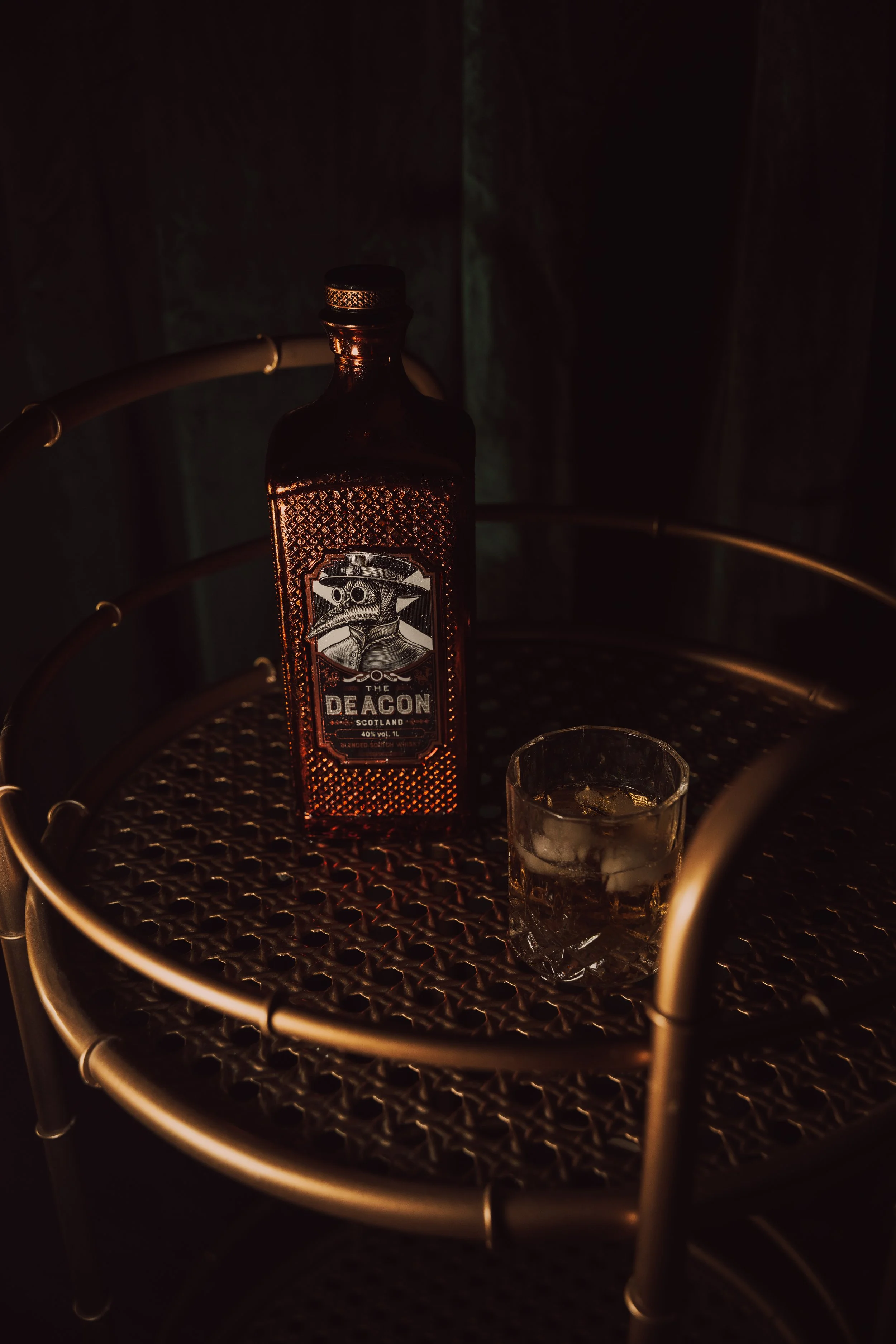 Whisky bottle and a tumbler of ice on a brass bistro table in a dark room, lit from one side