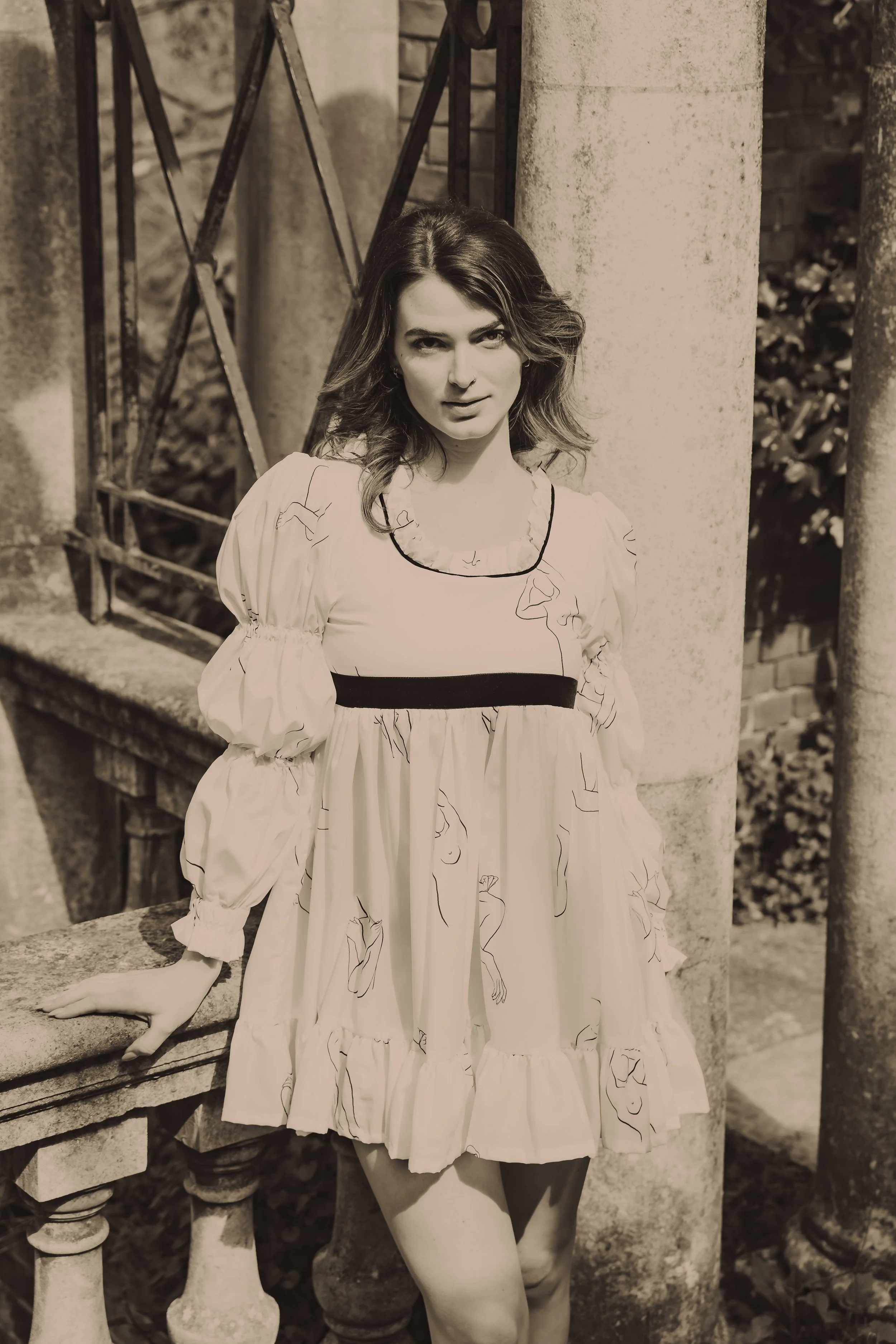 Black and white photo of a woman standing outdoors, leaning against a stone pillar next to a decorative railing, wearing a long, flowing dress with a ruffled neckline and puffed sleeves.