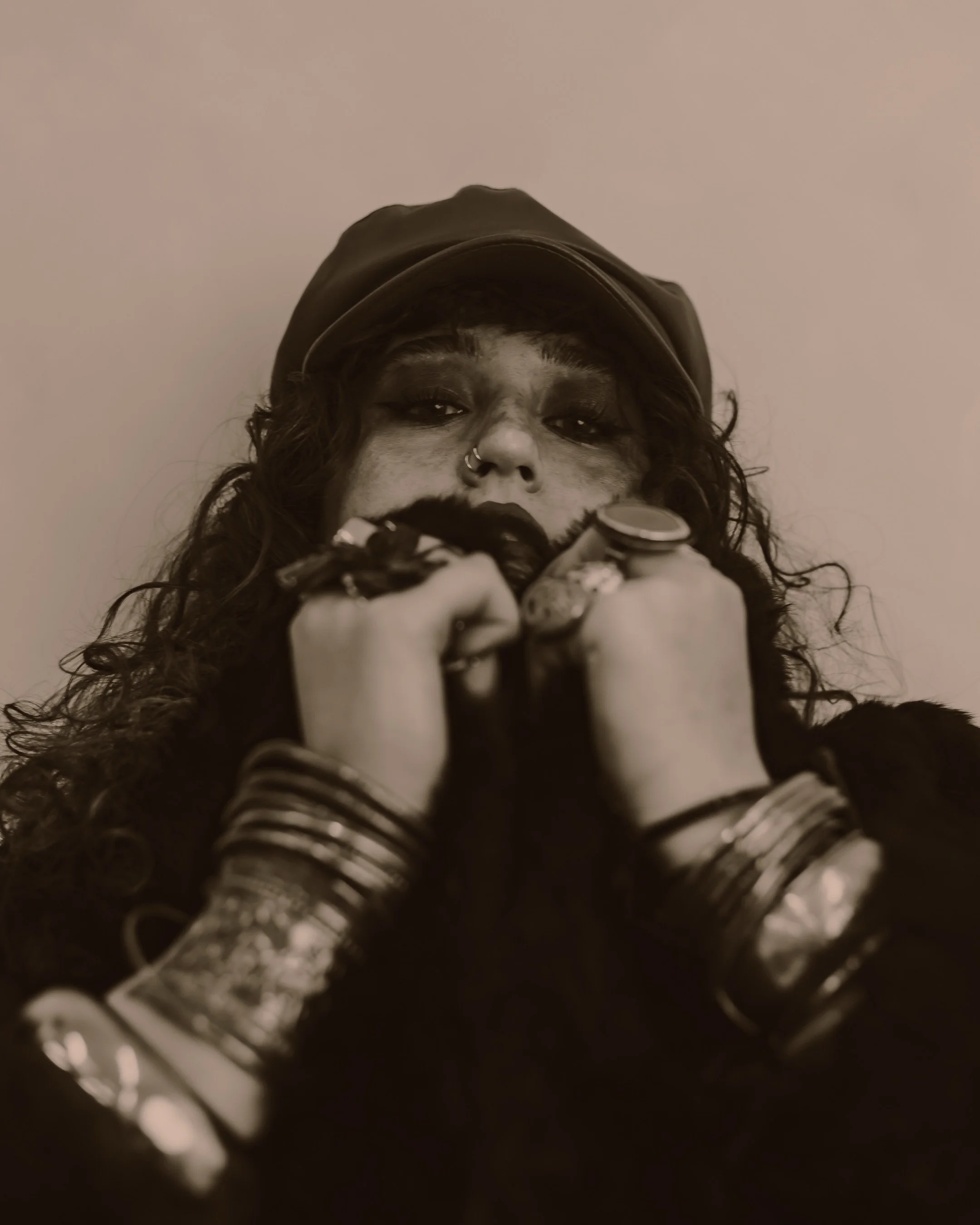 Close portrait of a woman in a flat cap with stacked bangles and rings, hands raised to her face, warm monochrome