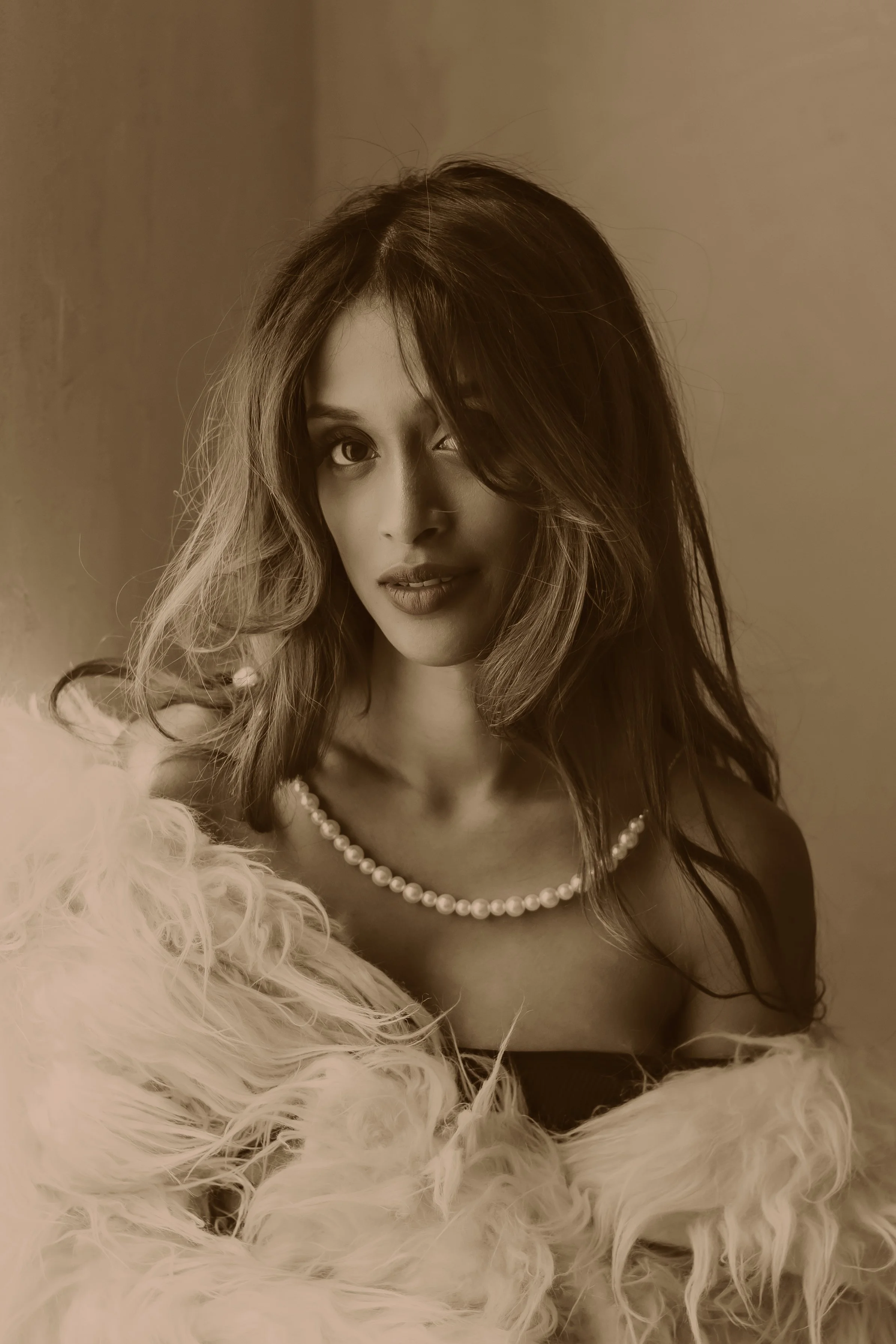 A woman with wavy hair wearing a string of pearls, a strapless top, and a fluffy fur coat, looking at the camera with a slight smile.