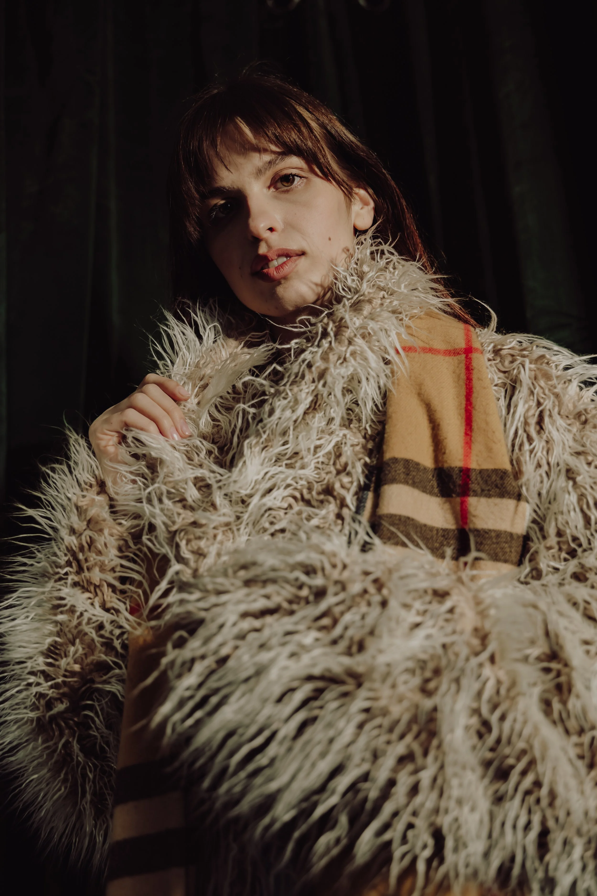 A woman with short brown hair wearing a fuzzy cream-colored coat and a Burberry-style scarf with a beige base and red and black plaid pattern, standing in front of a dark wooded background.