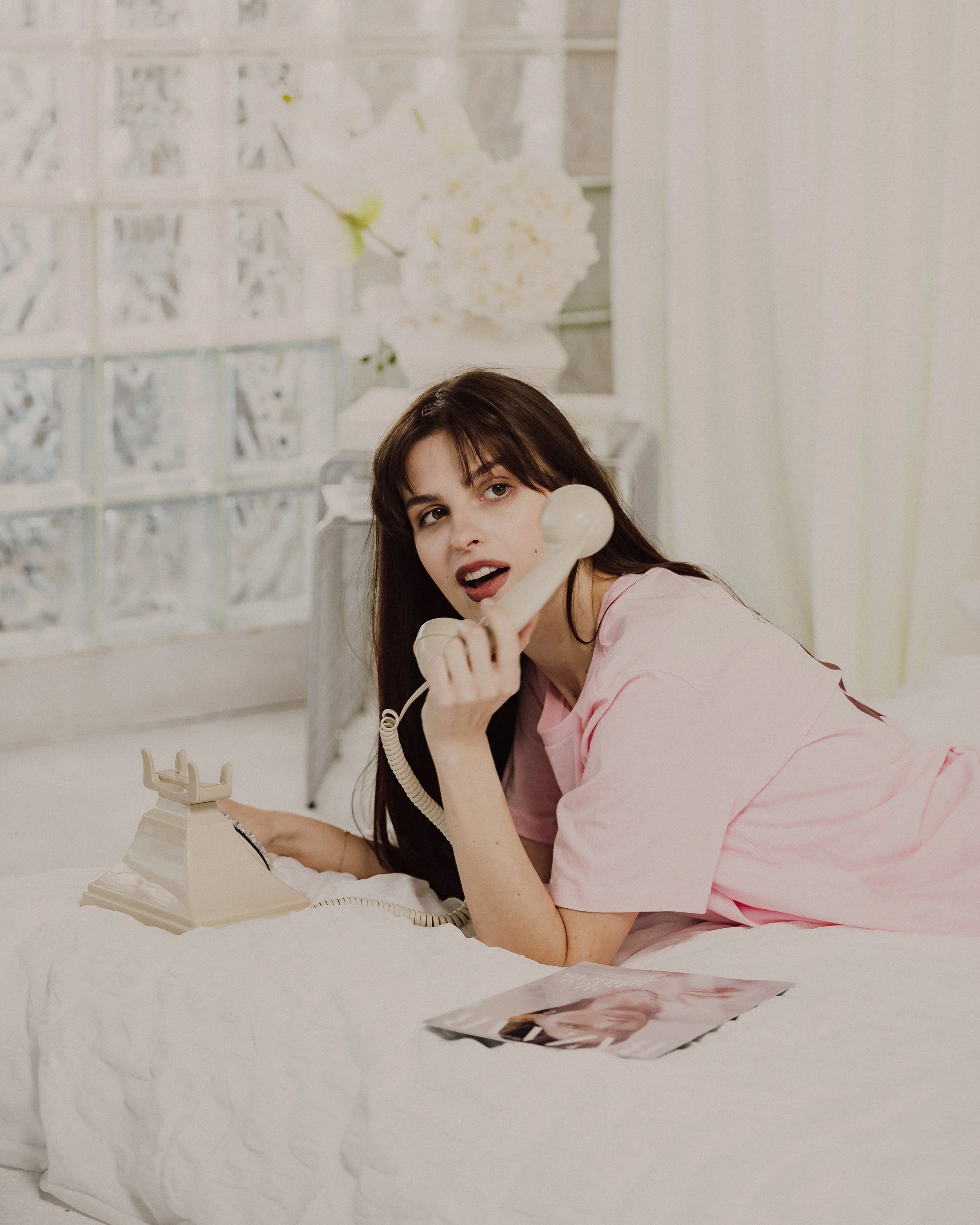 A young woman with long dark hair, wearing pink scrubs, talking on a beige rotary phone in a white room with a white blanket and a printed photograph of herself on the bed.