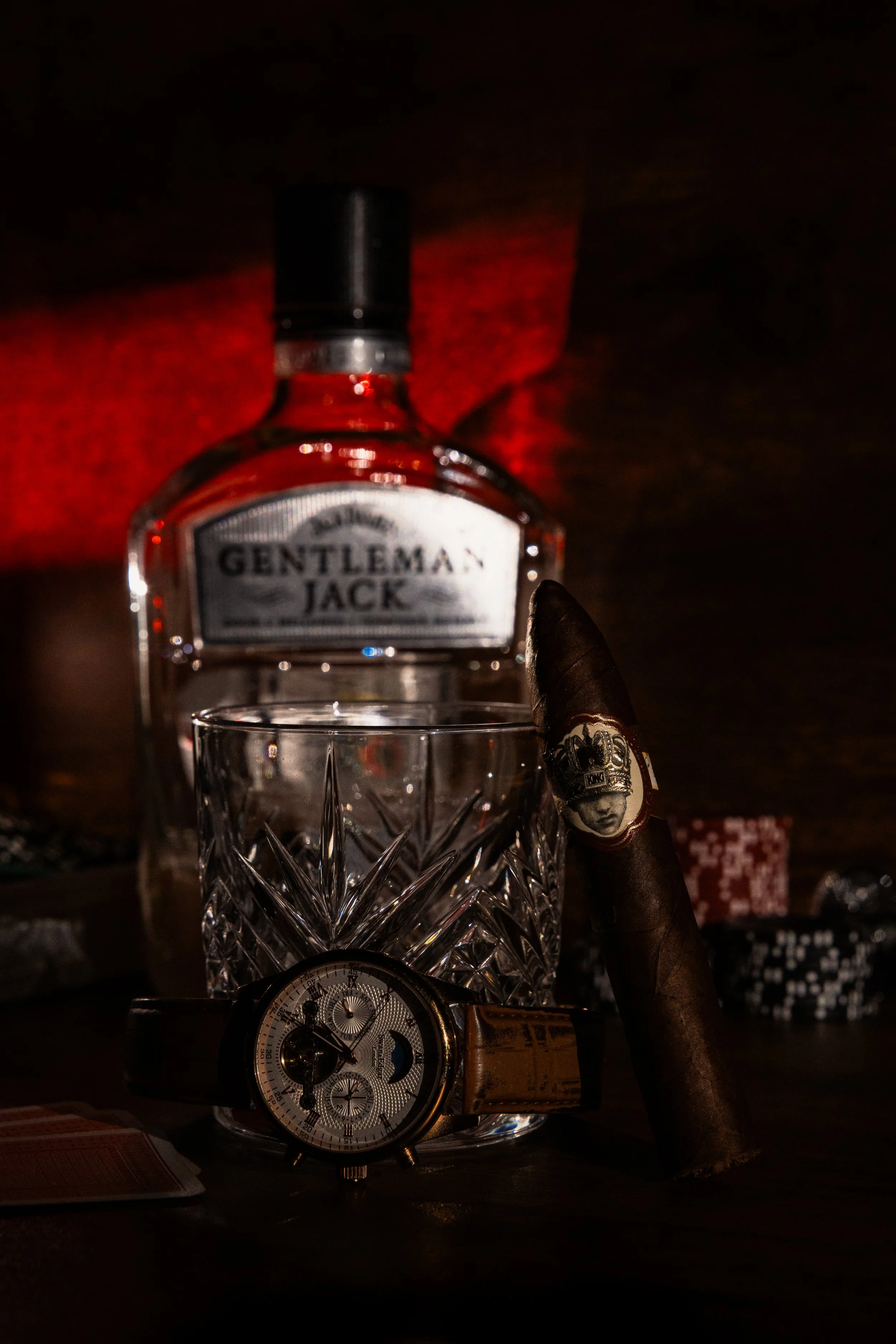 Cigar resting against a cut-crystal tumbler and a whiskey bottle under deep red light, with a gold watch and playing cards on a dark table