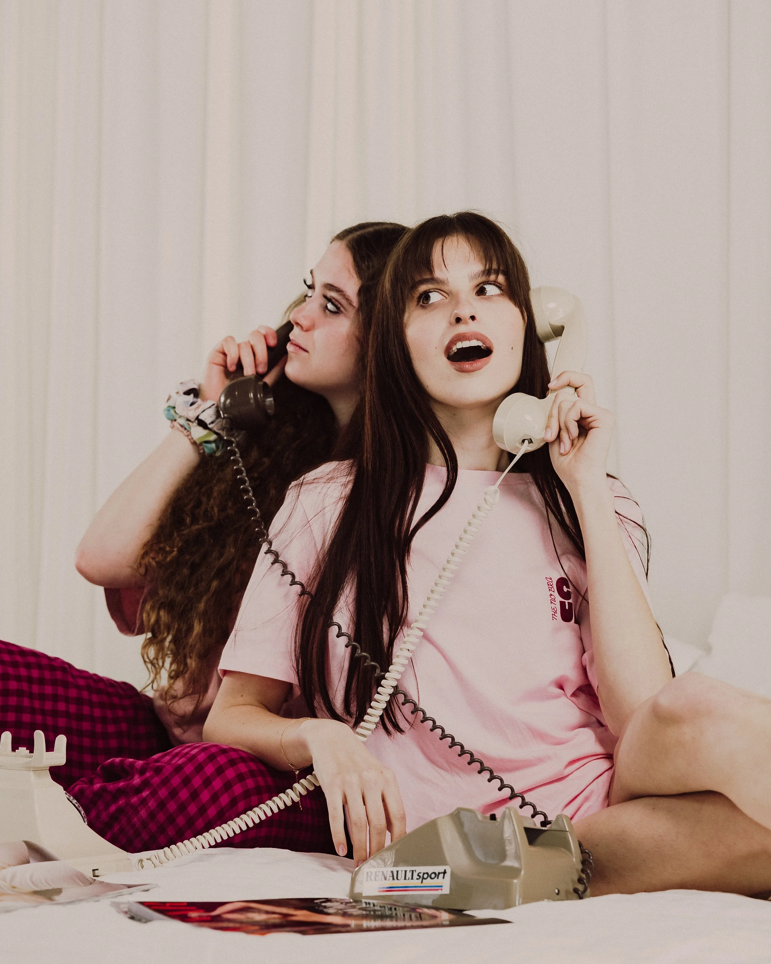 Two young women with long hair talking on vintage landline phones, with one on the left looking sideways and the other on the right looking more directly, sitting at a table with magazines and a calculator labeled 'RENAULTsport' in front of them.