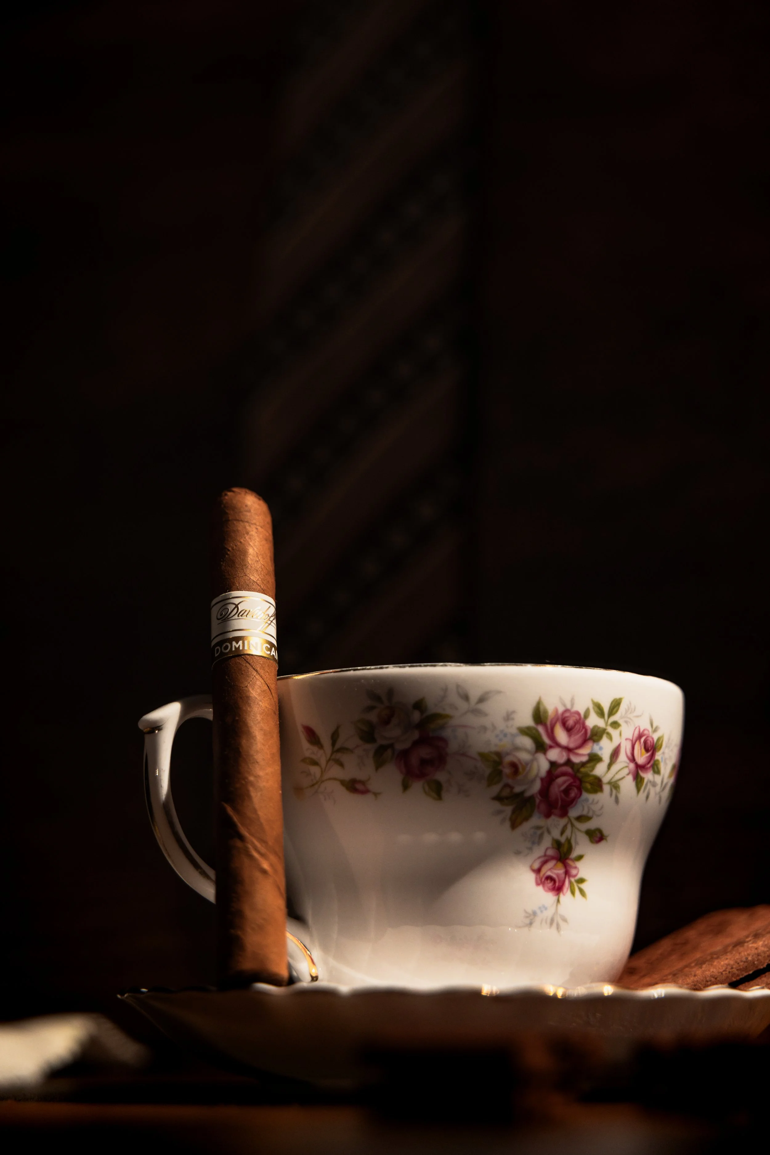 Hand-rolled cigar resting against a rose-patterned bone china teacup and saucer, warm low light